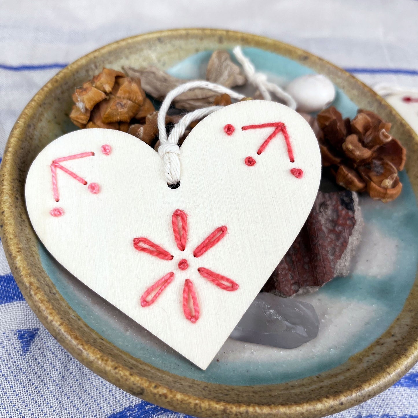 laser cut wood heart ornament embellished with hand embroidery in pink sitting on a dish of found nature objects on a cream and blue patterned scarf