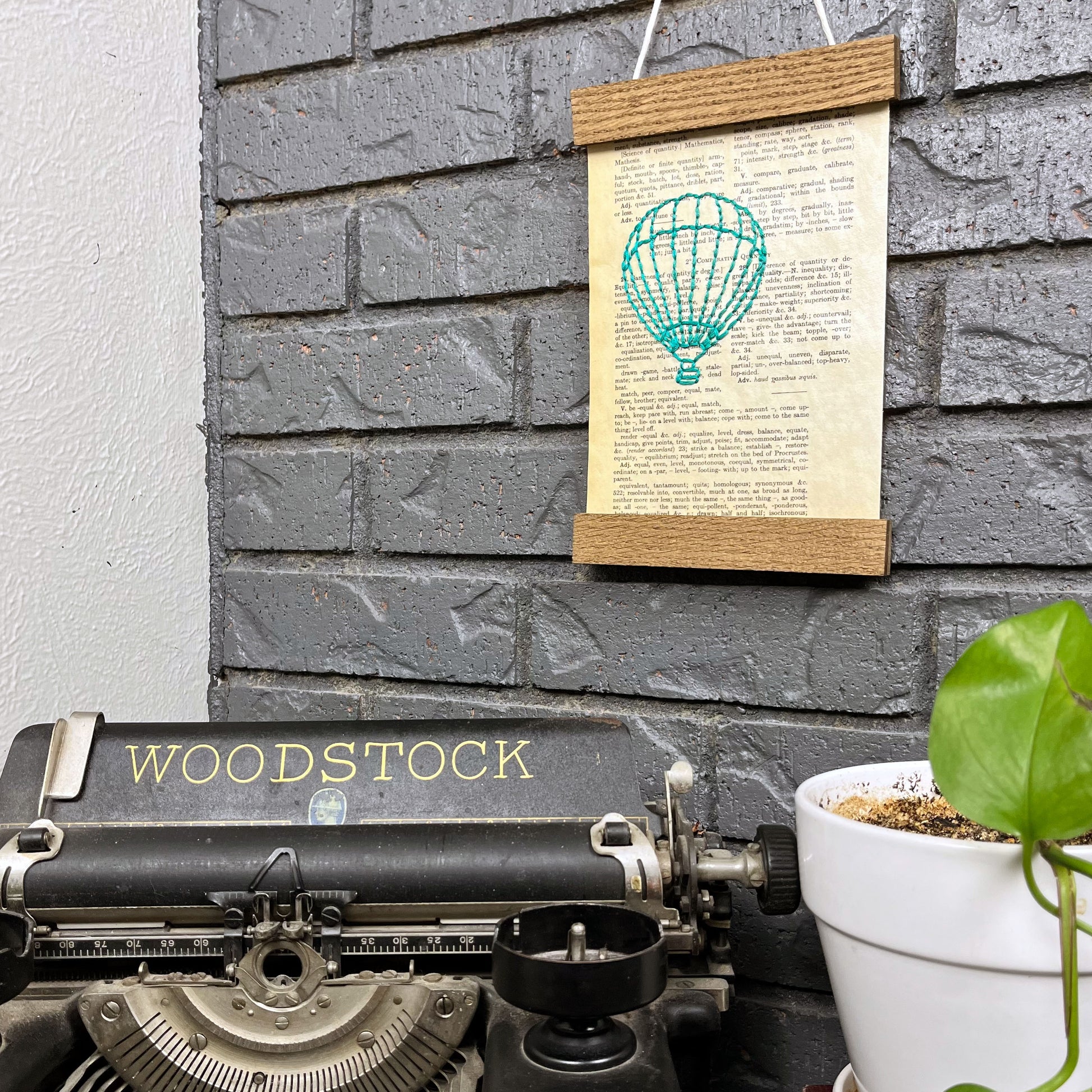 a vintage thesaurus page, hand stitched over with an outline of a hot air balloon in teal thread, hanging in a magnetic wood frame on a grey brick wall, above a typewriter and a pothos plant