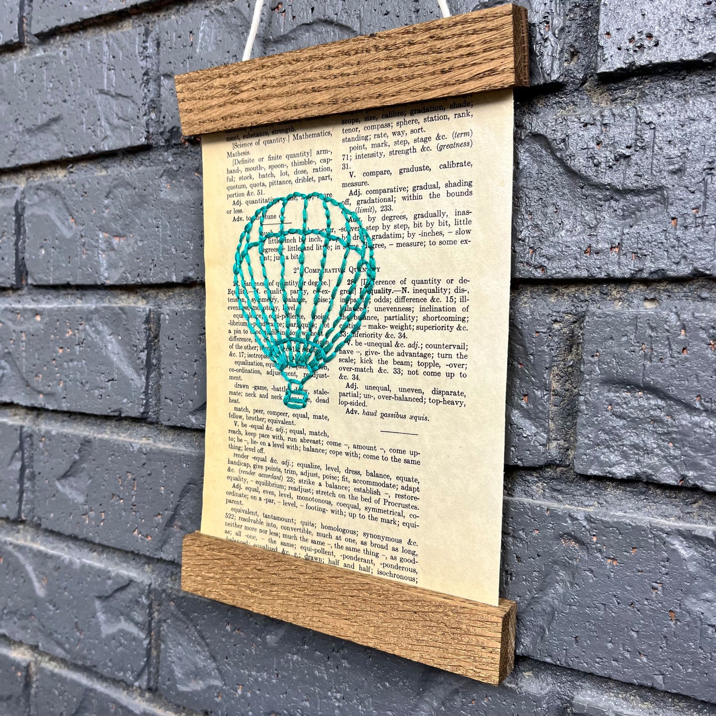 a vintage thesaurus page, hand stitched over with an outline of a hot air balloon in teal thread, hanging in a magnetic wood frame on a grey brick wall