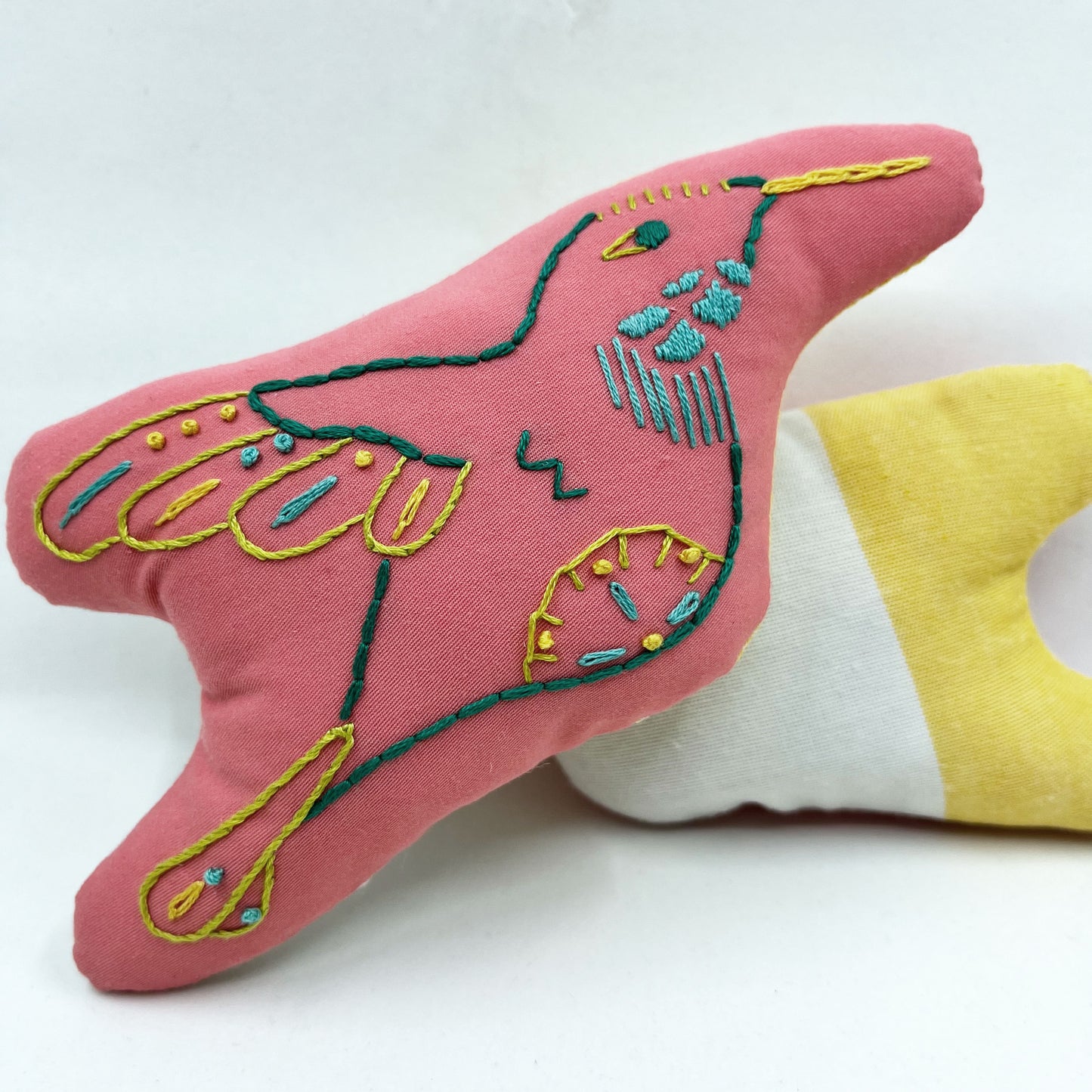 colorfully hand embroidered stuffed pillow animal of a hummingbird on coral fabric, behind it one turned around showing the back side made of shite and yellow color blocked fabric
