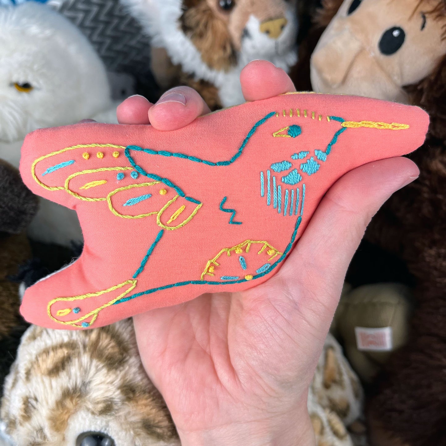 a hand holding a colorfully hand embroidered coral hummingbird, with other stuffed animals in the background