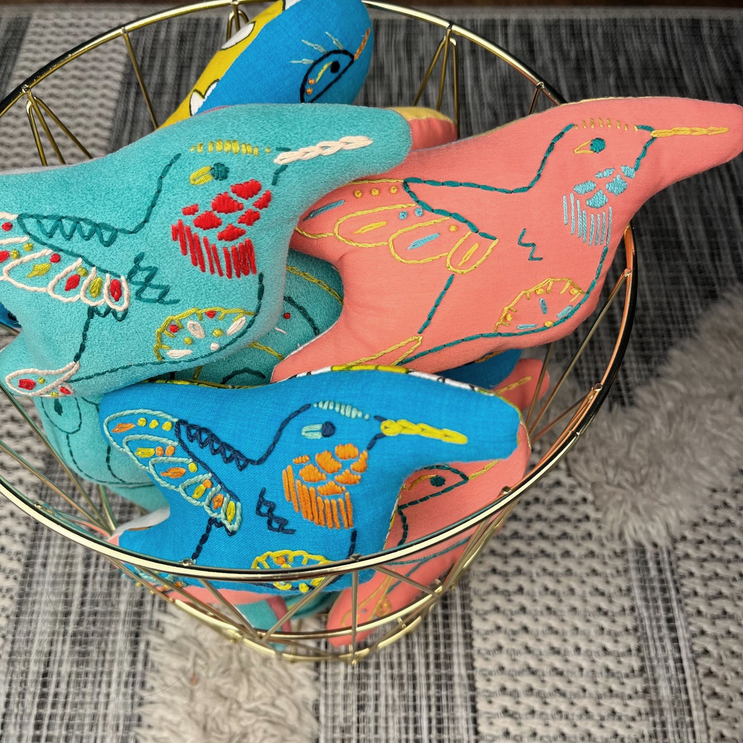 a group of colorfully hand embroidered stuffed pillow hummingbirds in blue, aqua and coral fabric, in a gold wire basket on a textured grey and white rug