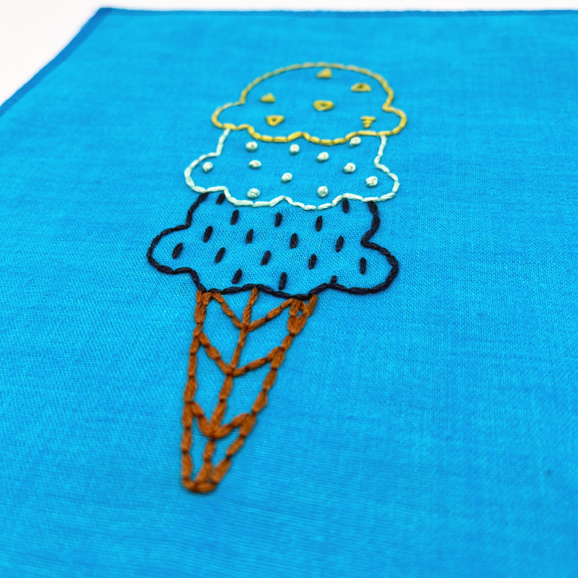 close up angled view ofa small bright blue fabric wall hanging, hand embroidered with with scoops of ice cream on a cone, the top scoop outlined and filled with yellow triangles, the middle mint green french dots, and the bottom dark blue dashes
