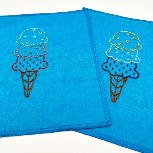 Two small wall hangings made out of bright blue woven fabric, hand embroidered ice cream cones, the one on the left in red coral and green, and the one on the right in yellow mint green and dark blue, each scoop is filled with different stitches