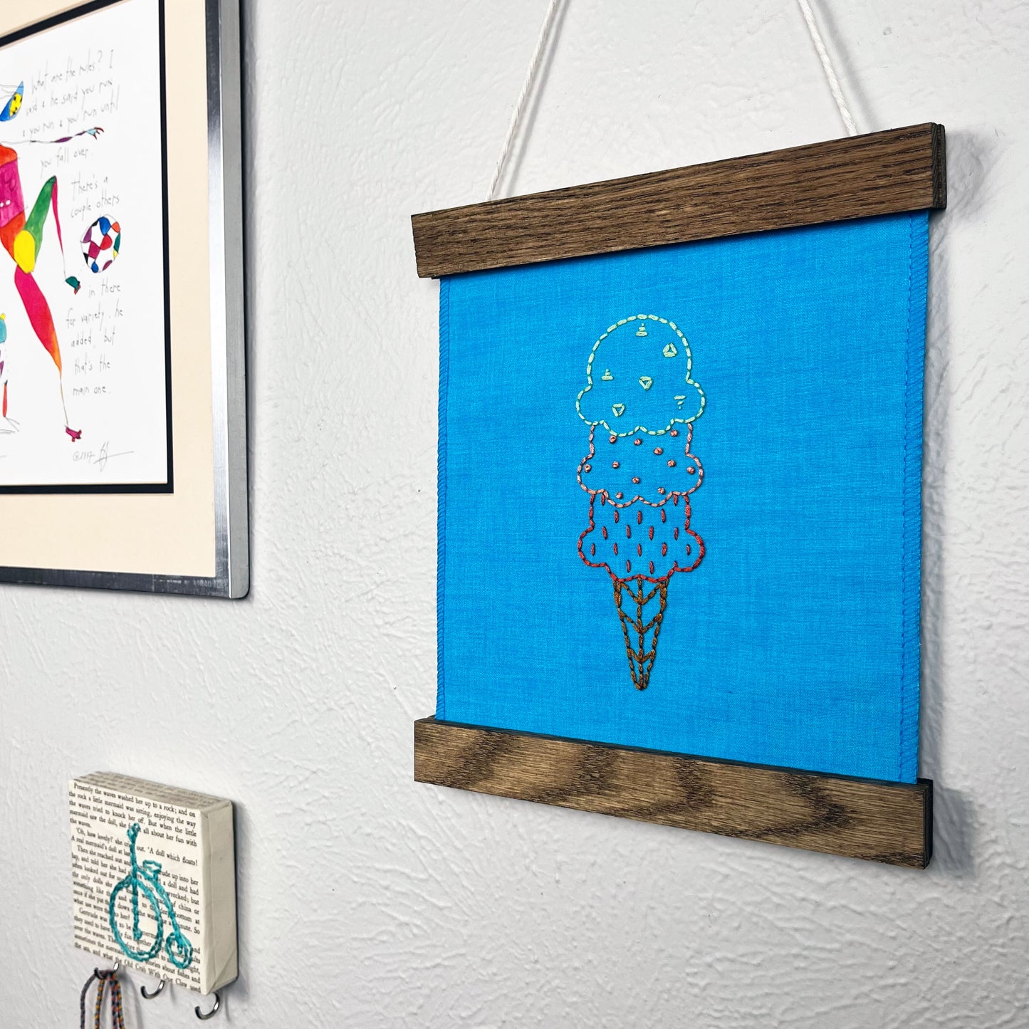 a small bright blue fabric wall hanging in a wood magnetic frame, hand embroidered with with scoops of ice cream on a cone, the top scoop outlined and filled with green triangles, the middle coral french dots, and the bottom red dashes