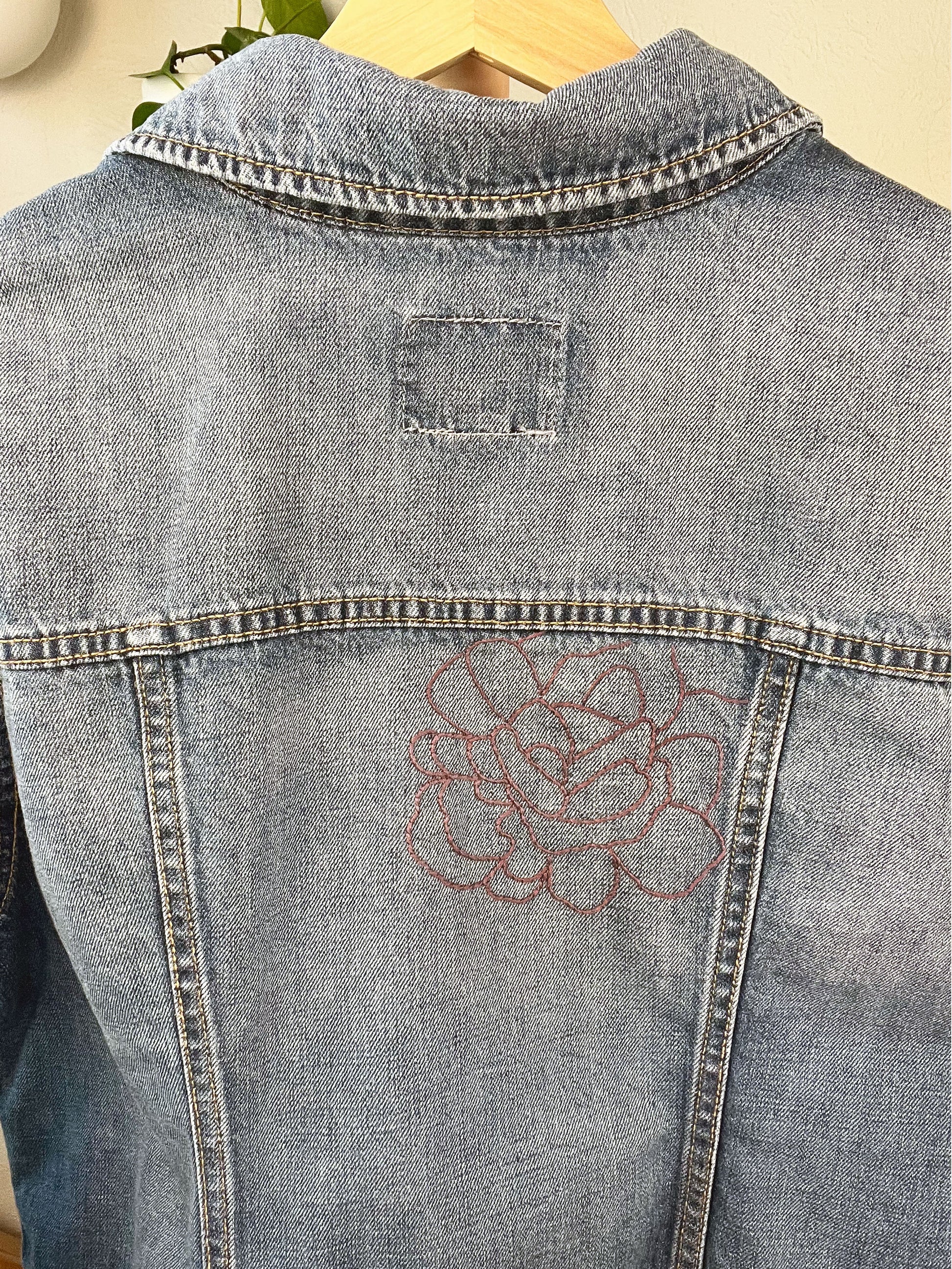back view of a denim jacket with an outline of a large peony off center in purple being held up on a hanger