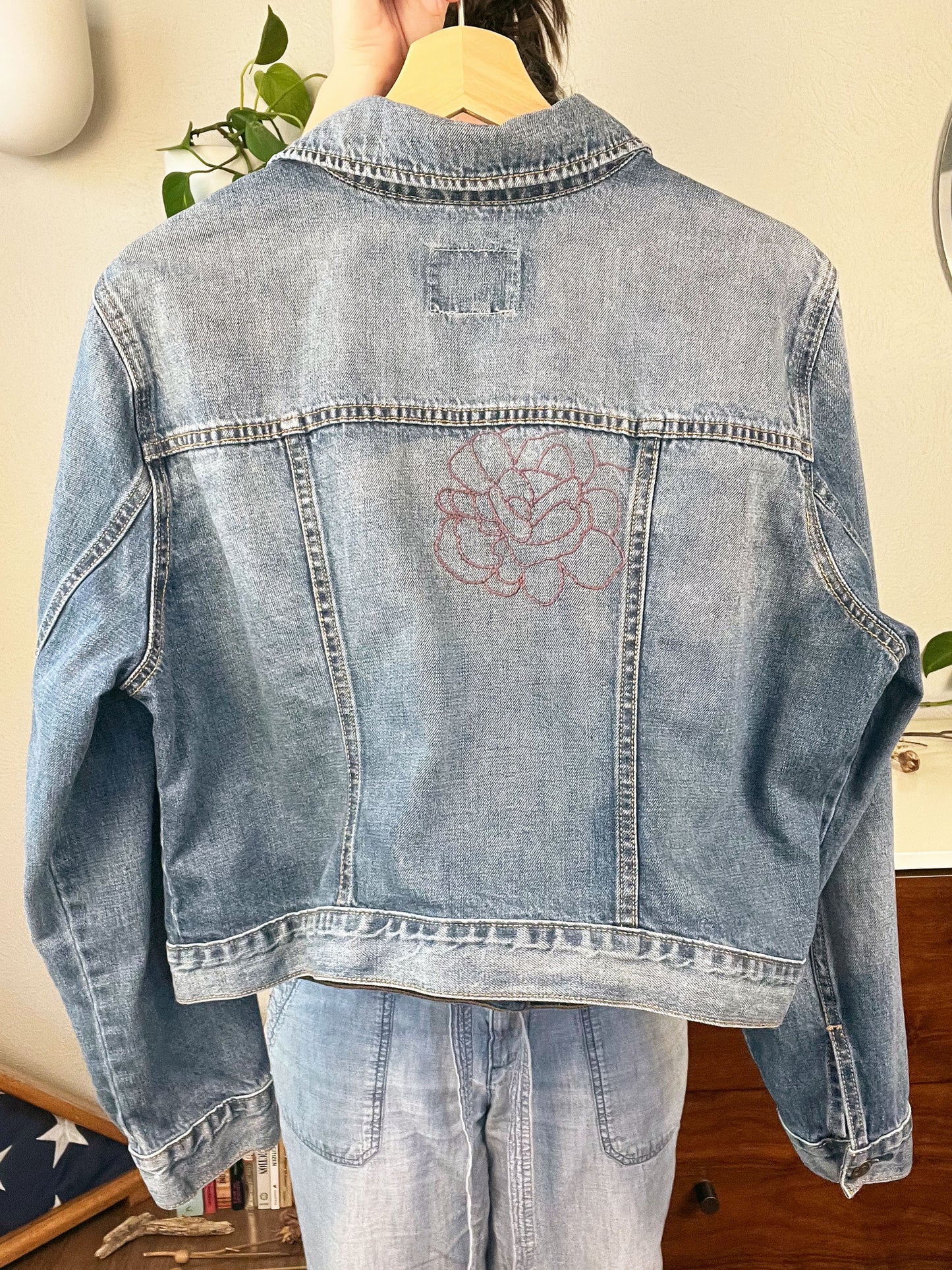 back view of a denim jacket with an outline of a large peony off center in purple being held up on a hanger