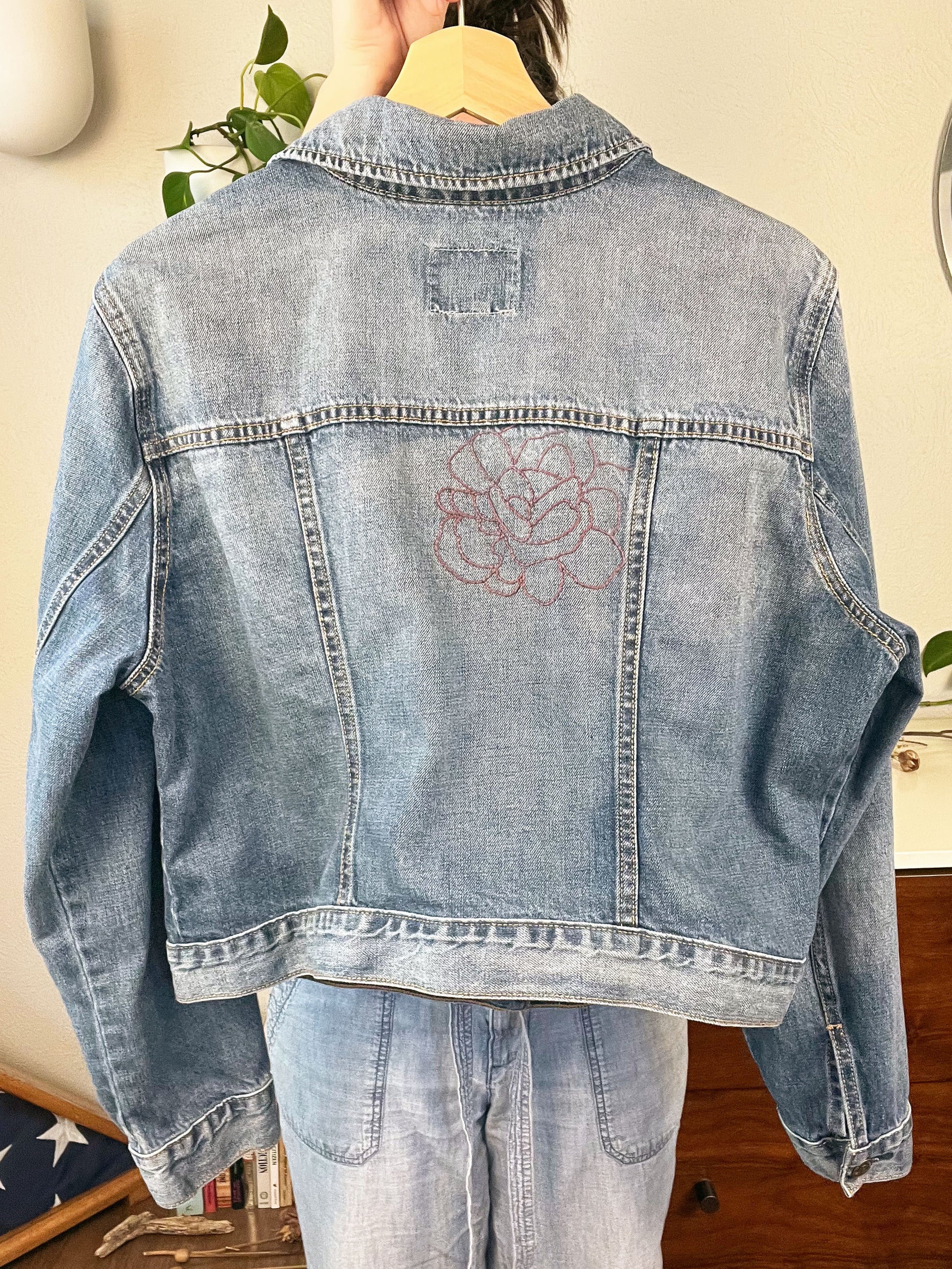 back view of a denim jacket with an outline of a large peony off center in purple being held up on a hanger