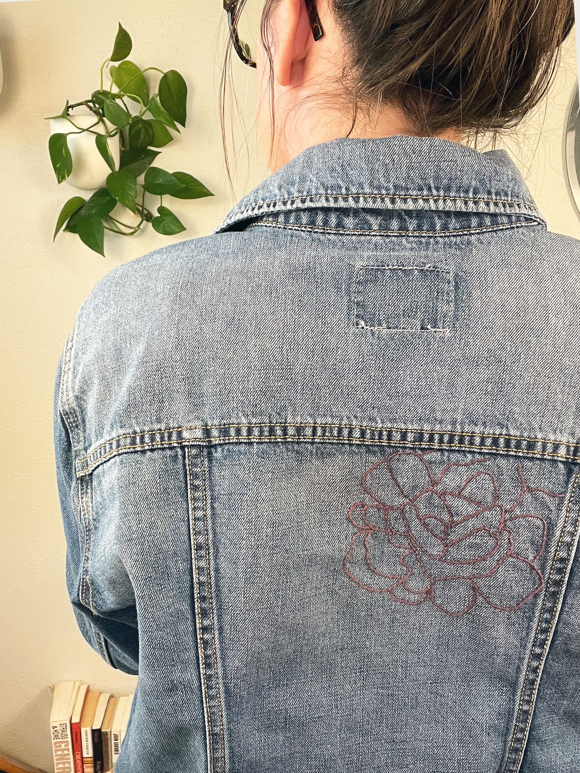 back view from the waist up of a woman wearing a denim jacket with an outline of a large peony off center in purple