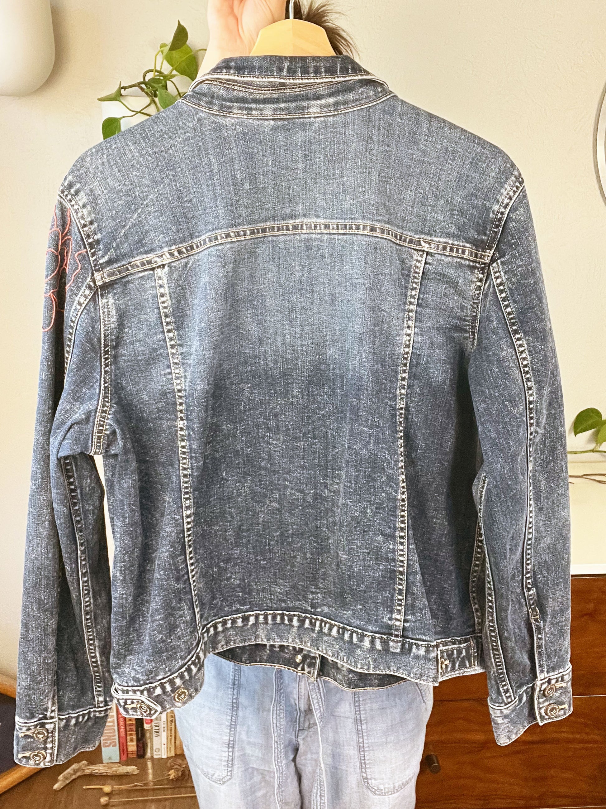 back view of a denim jacket being held up on a hanger, with a hand embroidered peony slightly visible on the left sleeve