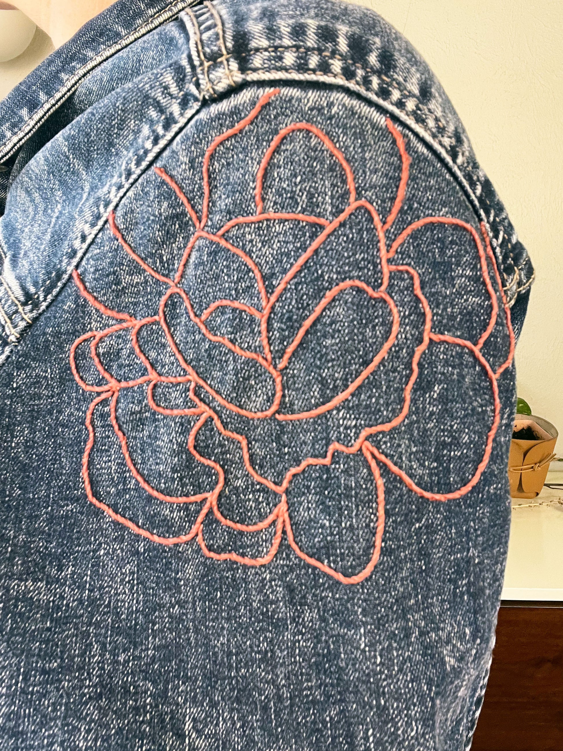 close up view of a hand embroidered outline of a large pink peony on the bicep of a denim jacket sleeve