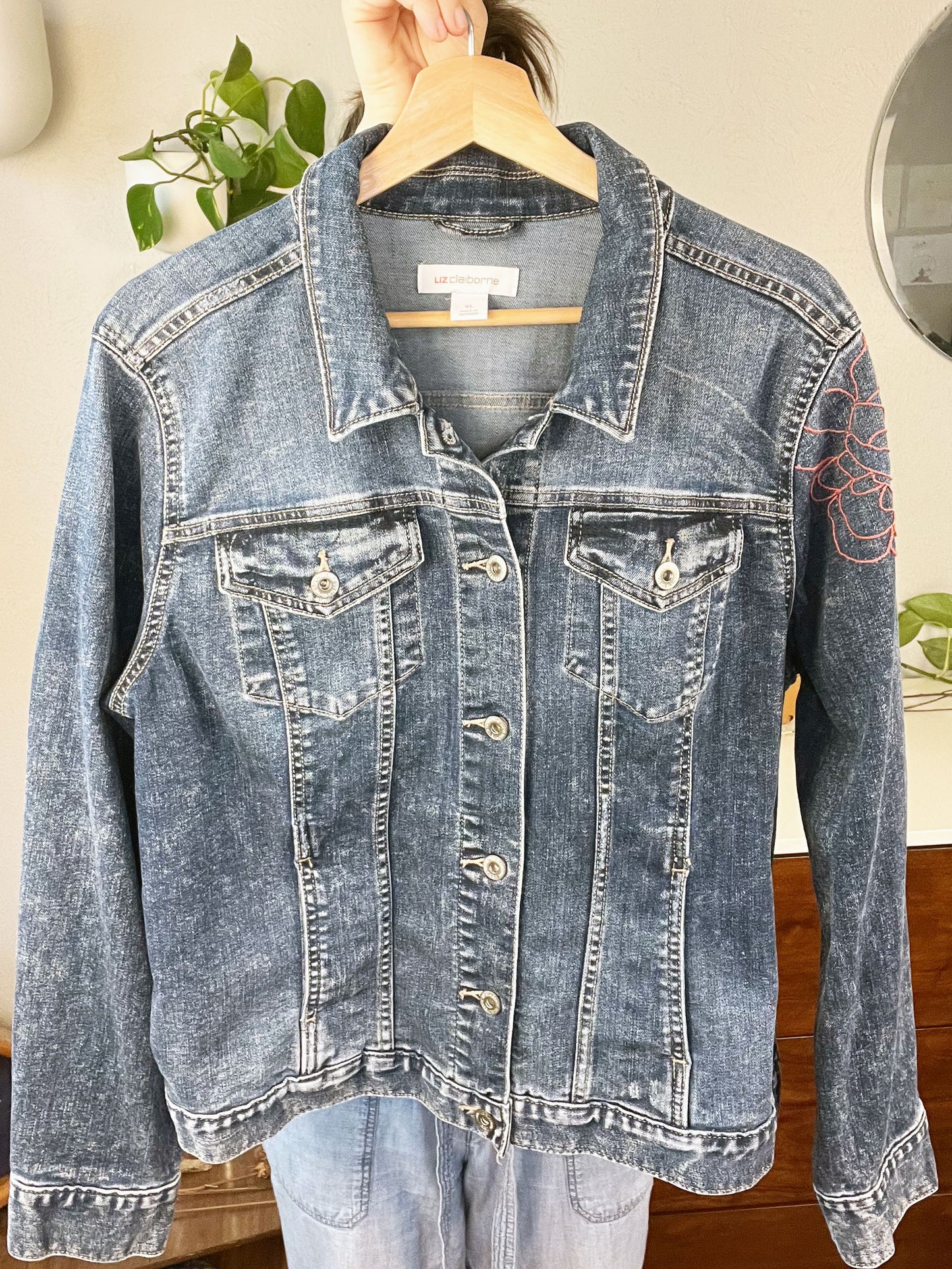 a denim jacket being held up on a hanger, with a hand embroidered peony slightly visible on the left sleeve