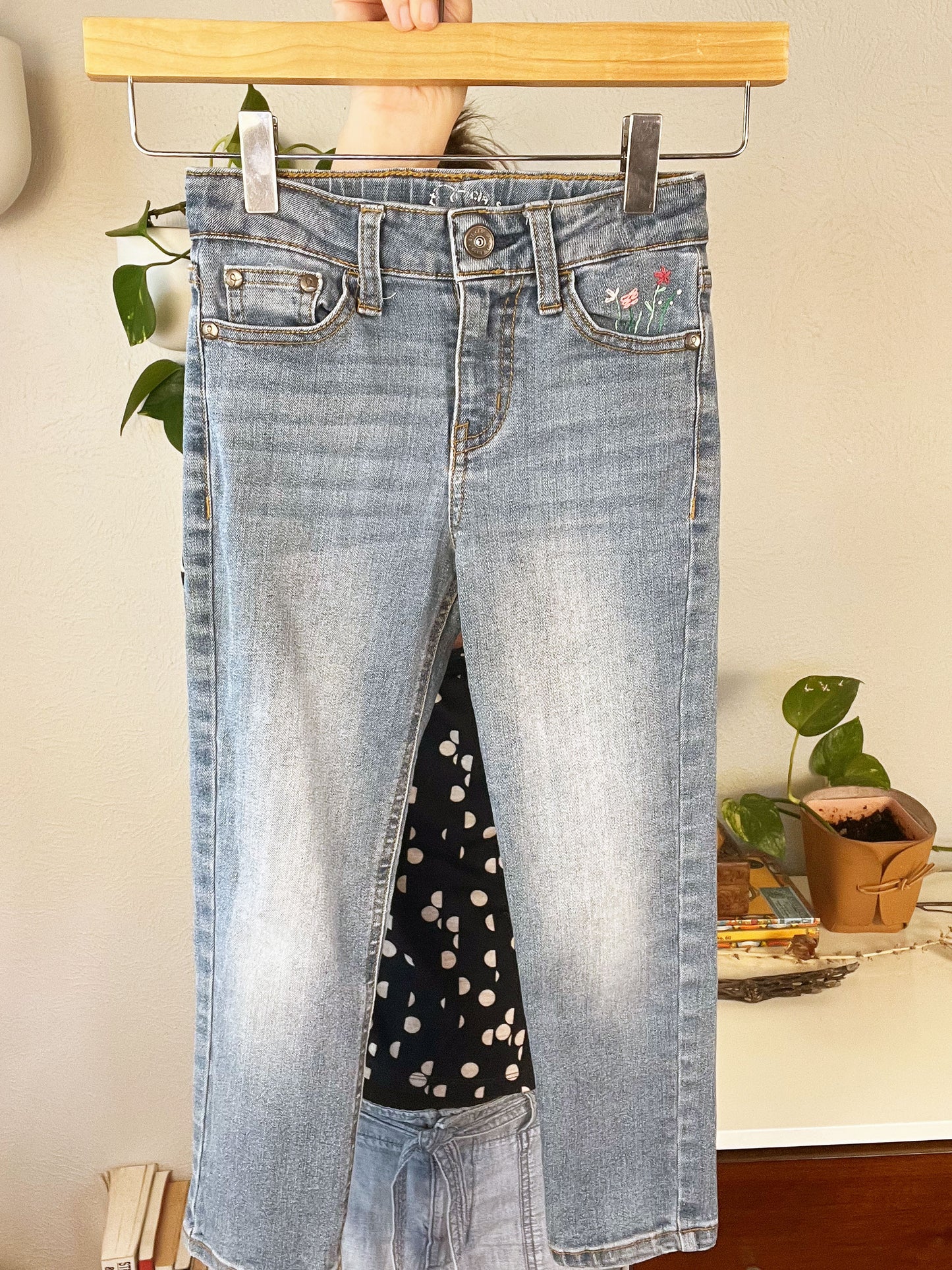 front view of blue jeans front pocket with hand embroidered flowers in greens and pinks, using chainstitch, stem stitch, and french knots, being held up on a hanger