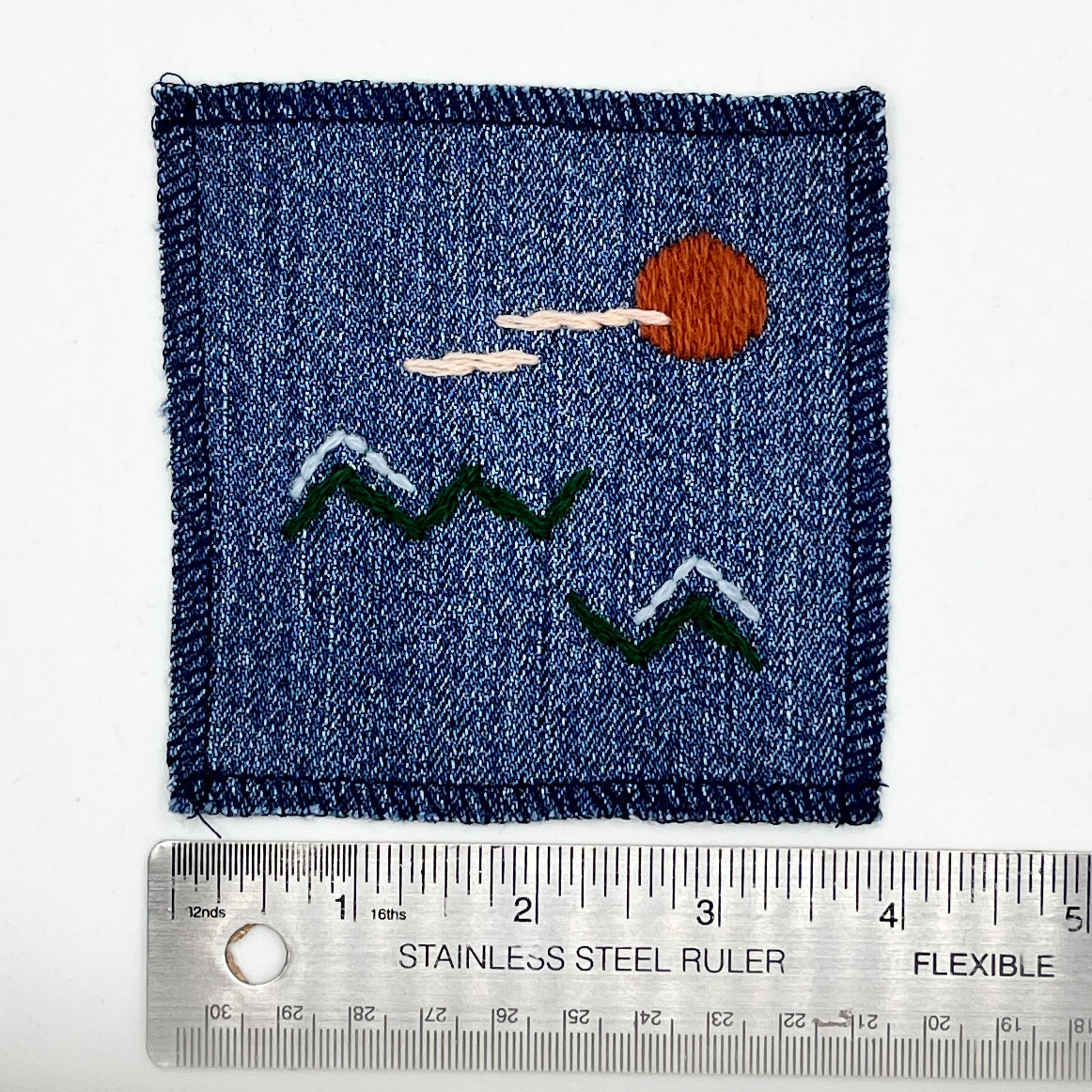 a square denim patch, with an abstract image hand stitched on them of a rust sun, with lines of peach clouds, over pine green zigzag mountains, some with a line of light blue snow on them, overlocked edges, next to a ruler showing a width of 4 inches