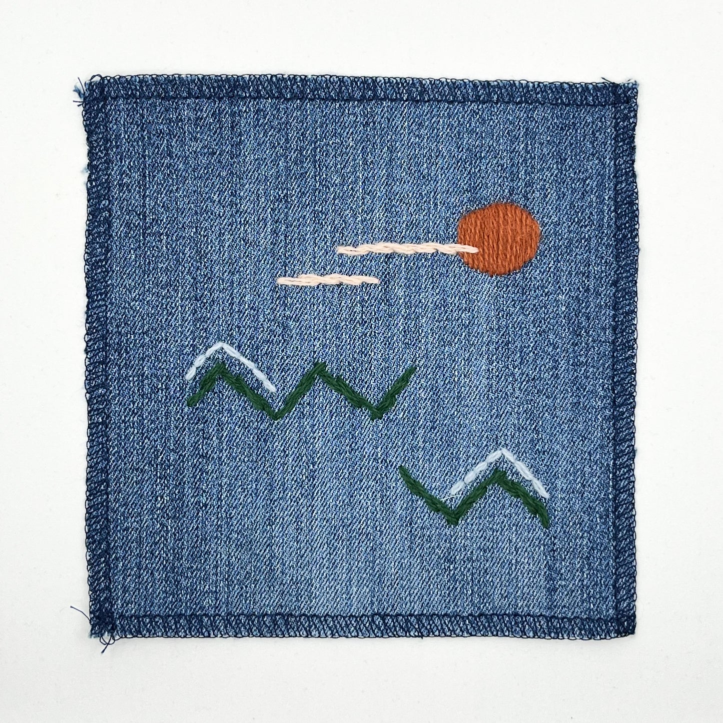 a square denim patch, with an abstract image hand stitched on them of a rust colored sun, with lines of peach clouds in front of it, over pine green zigzags as mountains, some with a line of light blue snow over them, overlocked edges