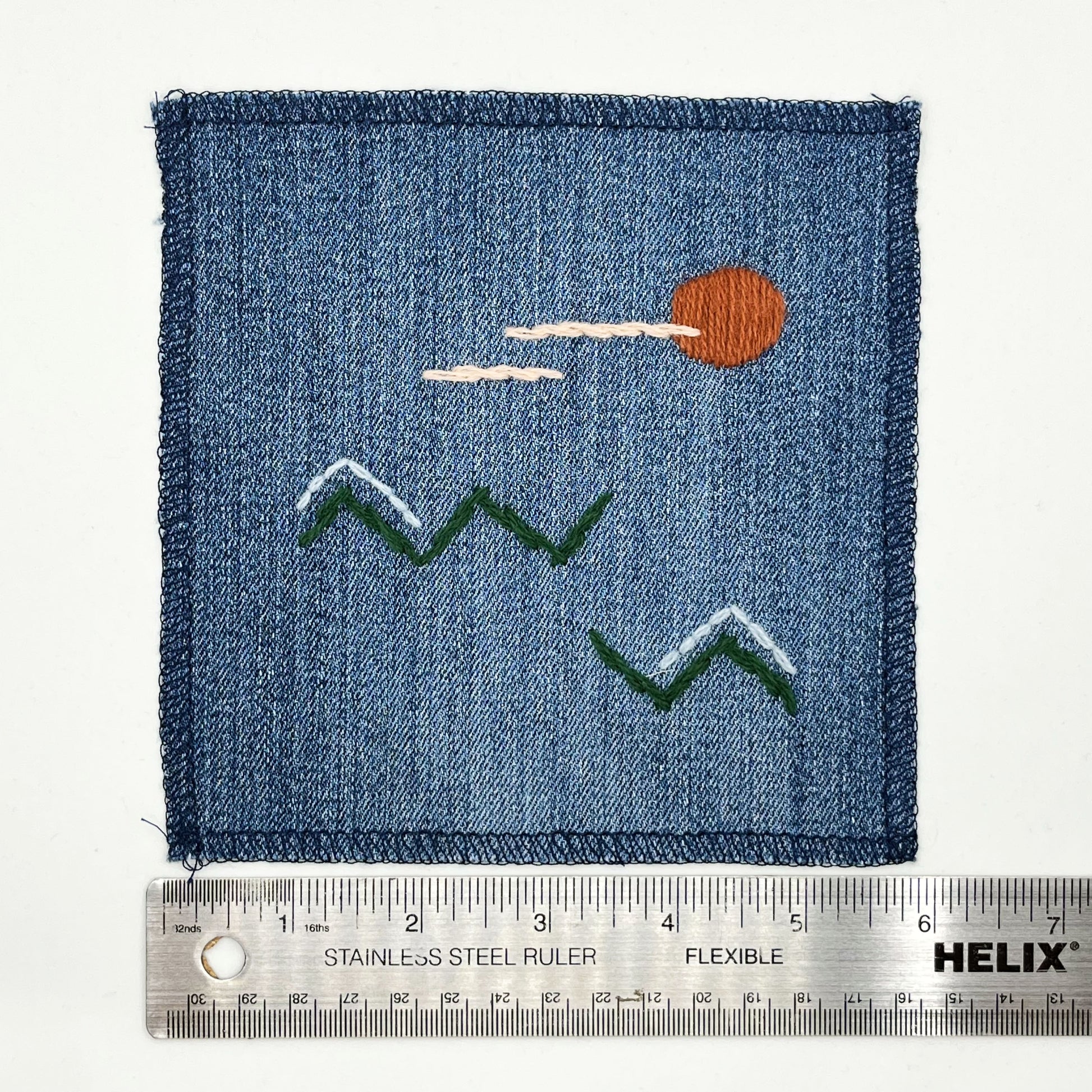a square denim patch, with an abstract image hand stitched on them of a rust sun, with lines of peach clouds, over pine green zigzag mountains, some with a line of light blue snow on them, overlocked edges, next to a ruler showing a width of 6 inches