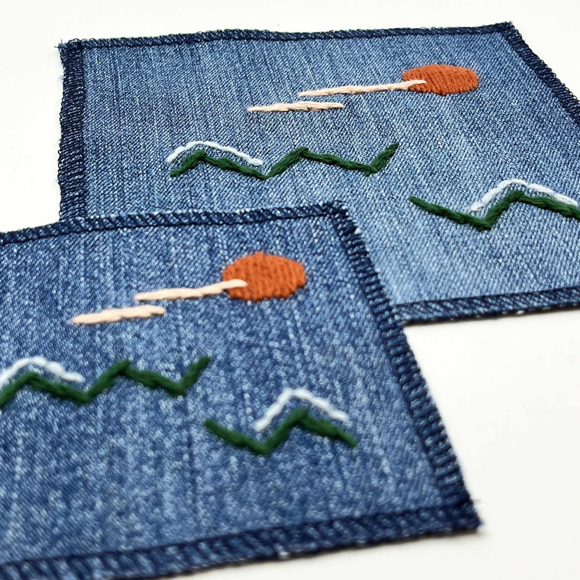 two square denim patches one small on medium, with an abstract image hand stitched on them of a rust colored sun, with lines of peach clouds in front of it, over pine green zigzags as mountains, some with a line of light blue snow over them