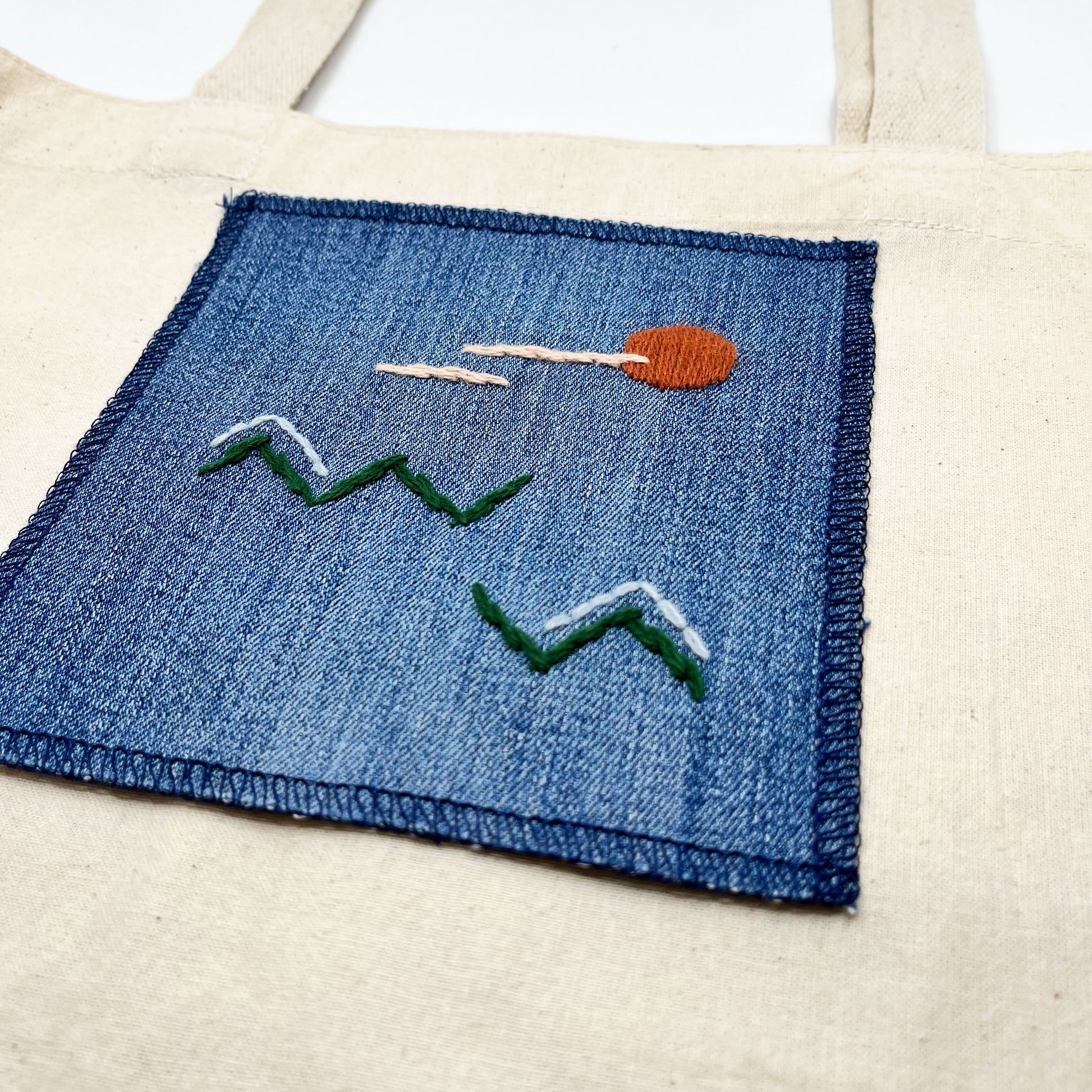 a square denim patch, with an abstract image hand stitched on them of a rust sun, with lines of peach clouds, over pine green zigzag mountains, some with a line of light blue snow on them, overlocked edges, on a canvas tote bag