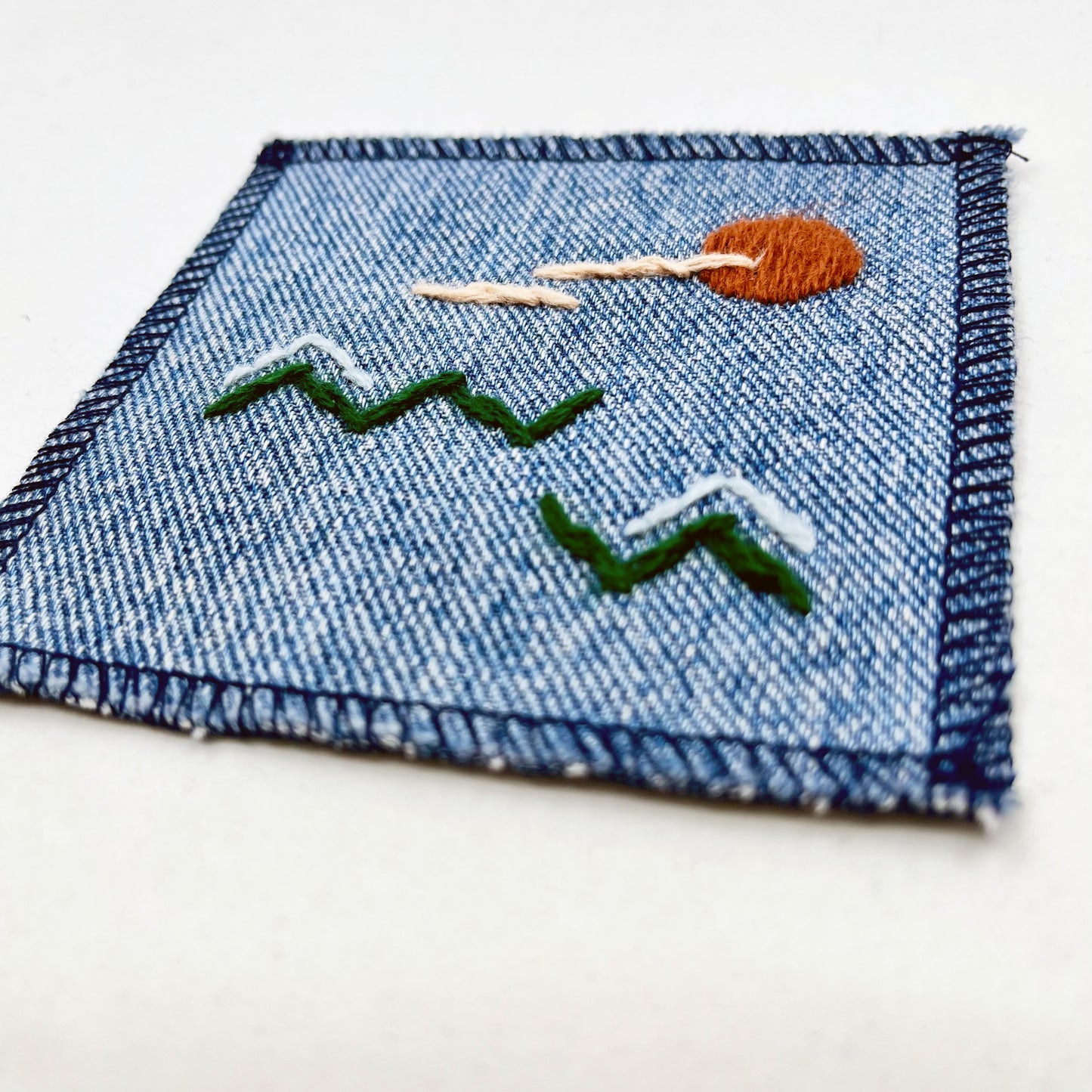 close up view of a square denim patch, with an abstract image handstitched on them of a rust colored sun, with lines of peach clouds in front of it, over pine green zigzags as mountains, some with a line of light blue snow over them, overlocked edges