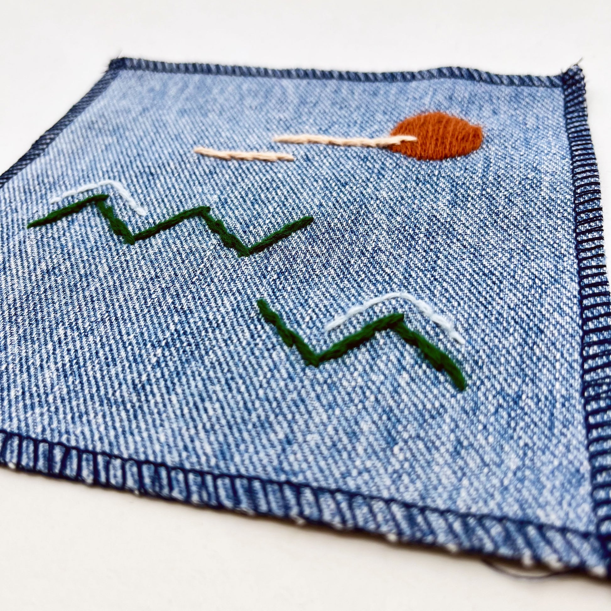 close up view of a square denim patch, with an abstract image handstitched on them of a rust colored sun, with lines of peach clouds in front of it, over pine green zigzags as mountains, some with a line of light blue snow over them, overlocked edges