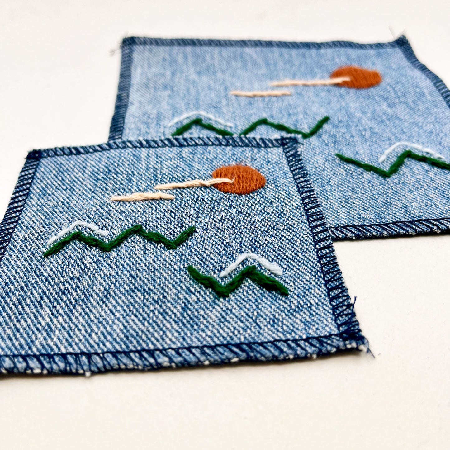 two square denim patches one small on medium, with an abstract image hand stitched on them of a rust colored sun, with lines of peach clouds in front of it, over pine green zigzags as mountains, some with a line of light blue snow over them