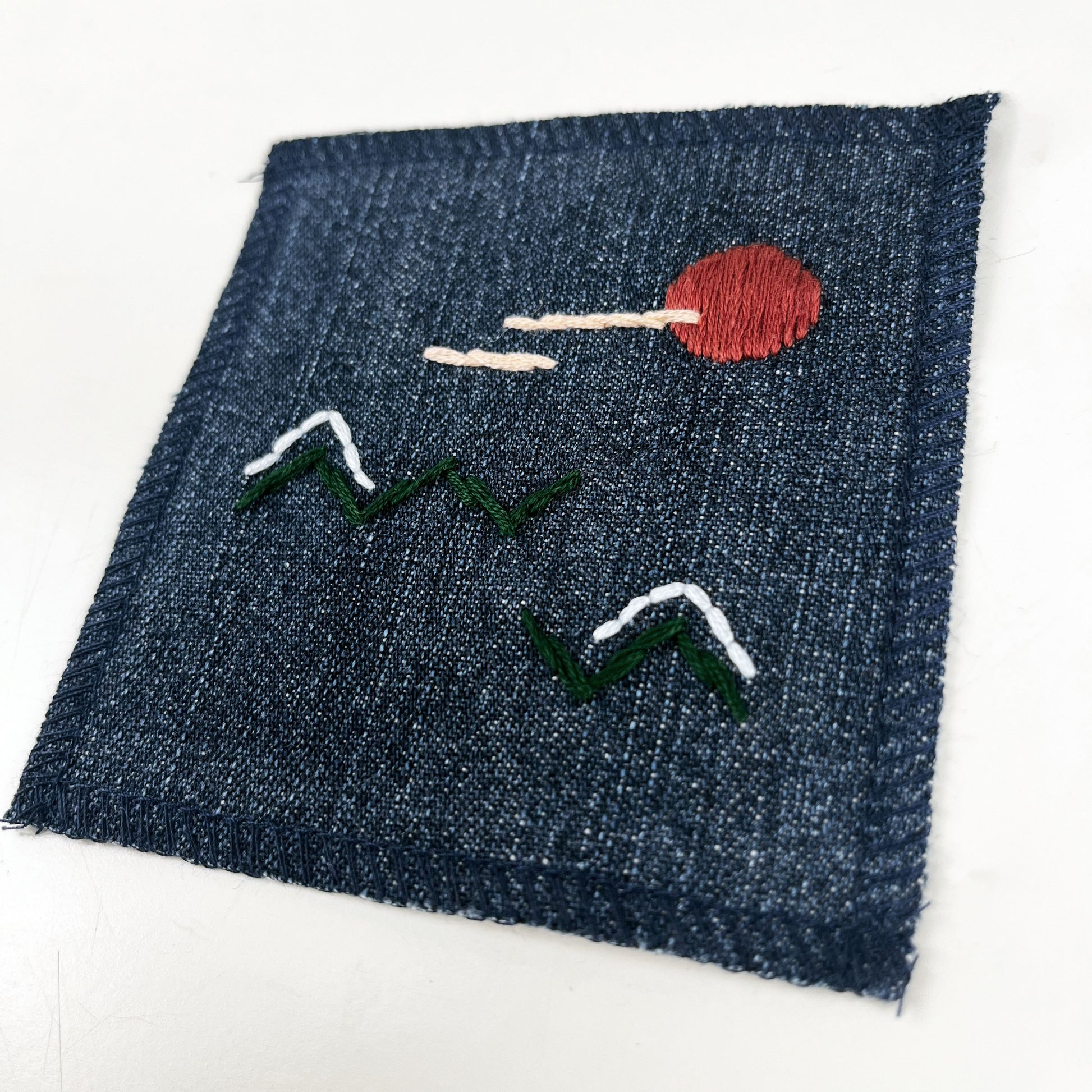 a square denim patch, with an abstract image hand stitched on them of a rust colored sun, with lines of peach clouds in front of it, over pine green zigzags as mountains, some with a line of light blue snow over them, overlocked edges
