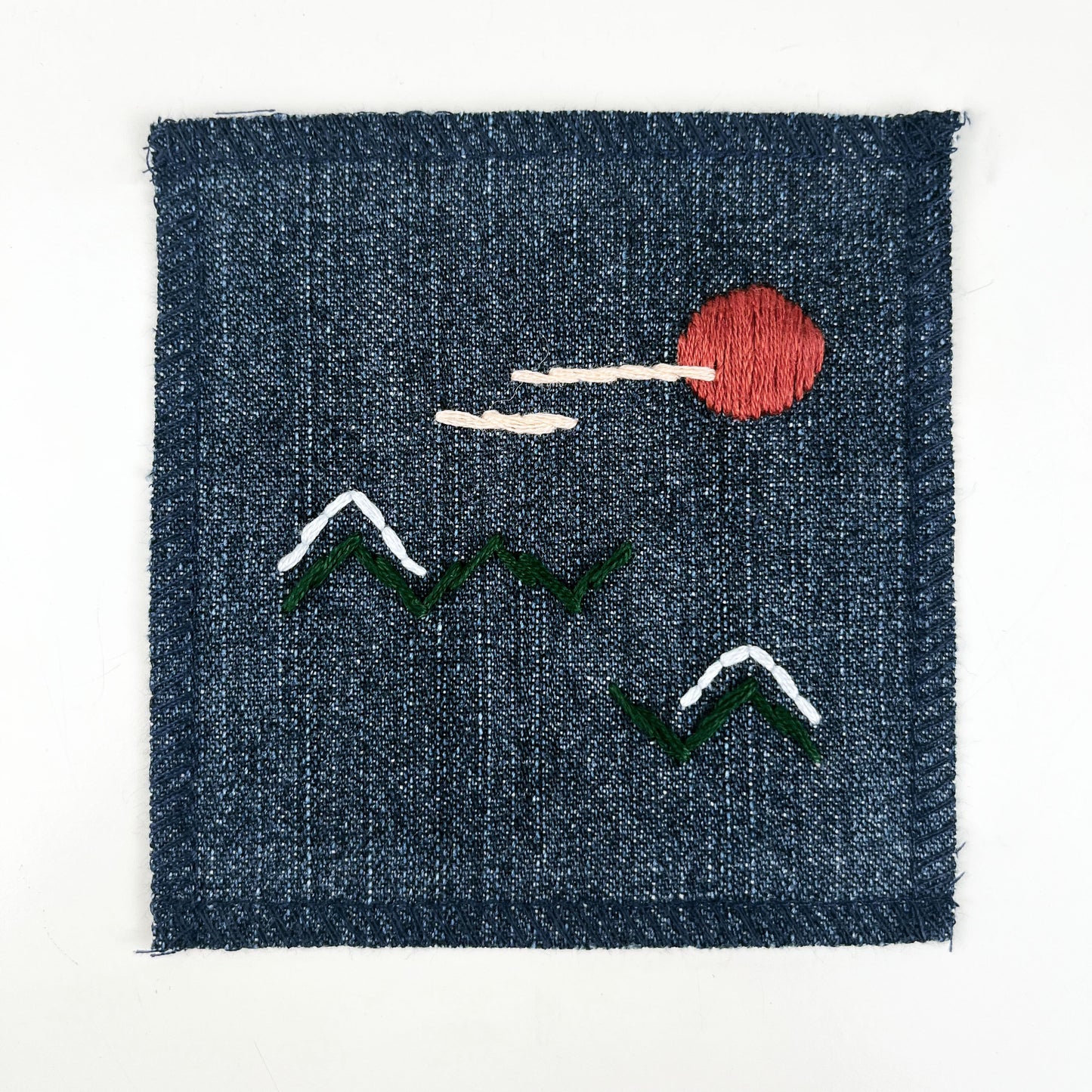 a square denim patch, with an abstract image hand stitched on them of a rust colored sun, with lines of peach clouds in front of it, over pine green zigzags as mountains, some with a line of light blue snow over them, overlocked edges