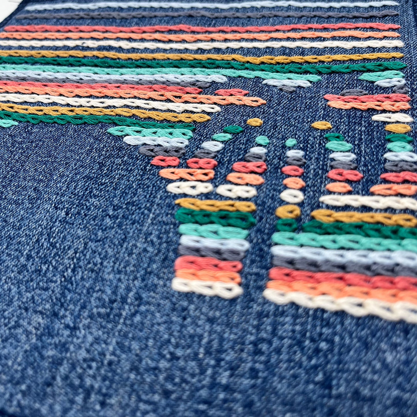 a close up angled view of a large square denim patch, hand stitched with rows of chainstitch, with negative space showing the shapes of a palm tree next to a setting sun, with peach, coral, grey, light blue, seafoam green and mustard colored thread
