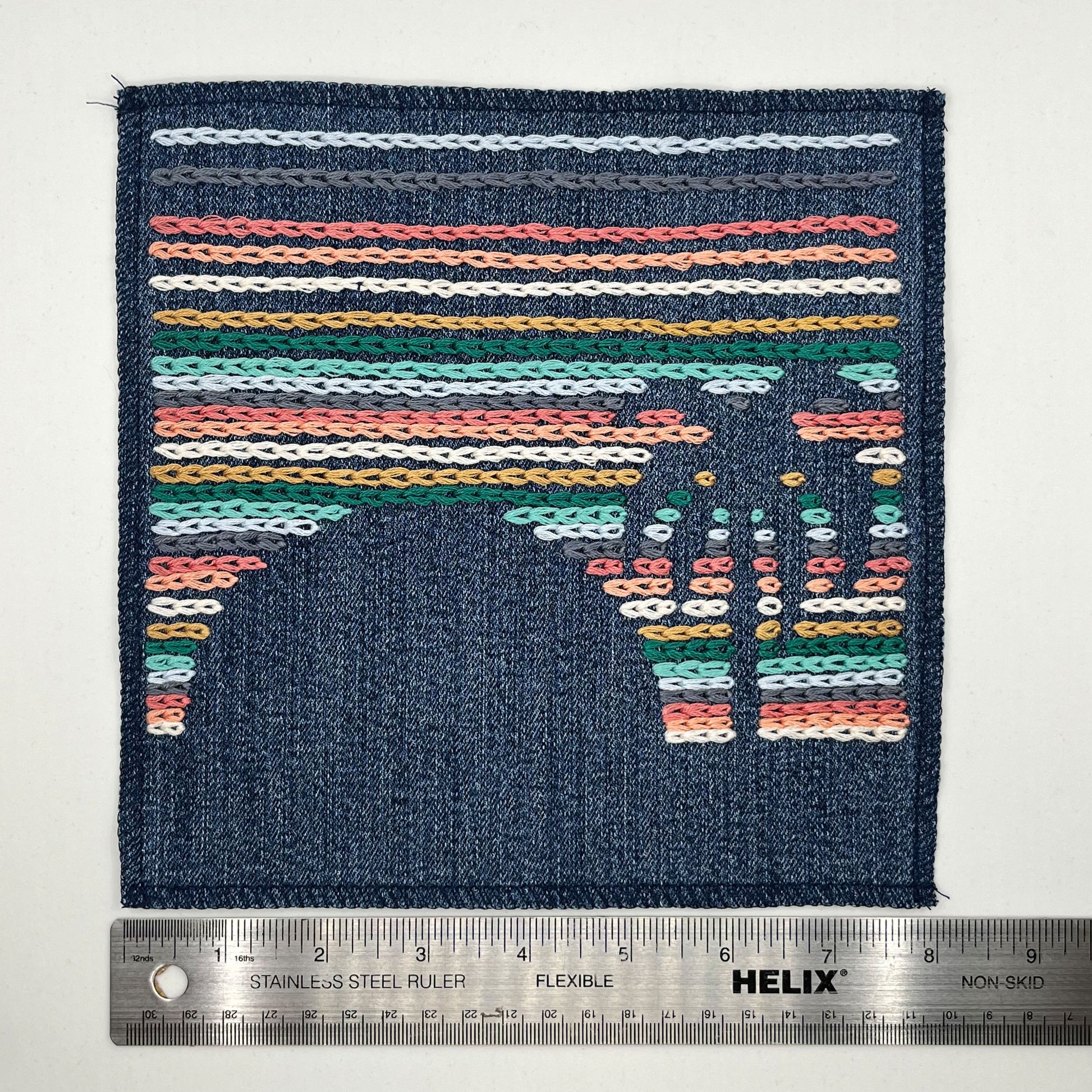 a large square denim patch, hand stitched with rows of chainstitch, with negative space showing the shapes of a palm tree next to a setting sun, with peach, coral, grey, light blue, green and mustard thread, next to a ruler showing a width of 8 in