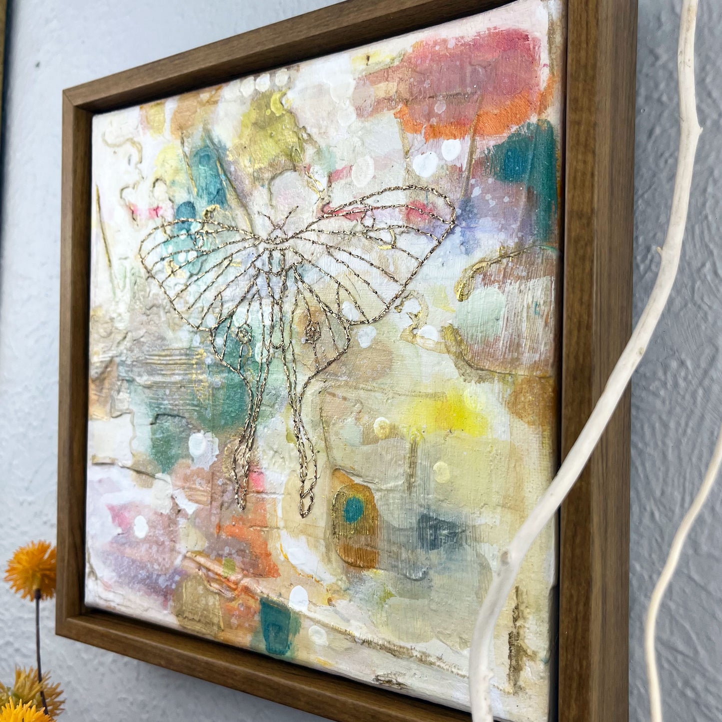 colorful square painting in a wood frame, with a line drawing of a luna moth hand stitched through it in metallic gold thread, hanging on a wall