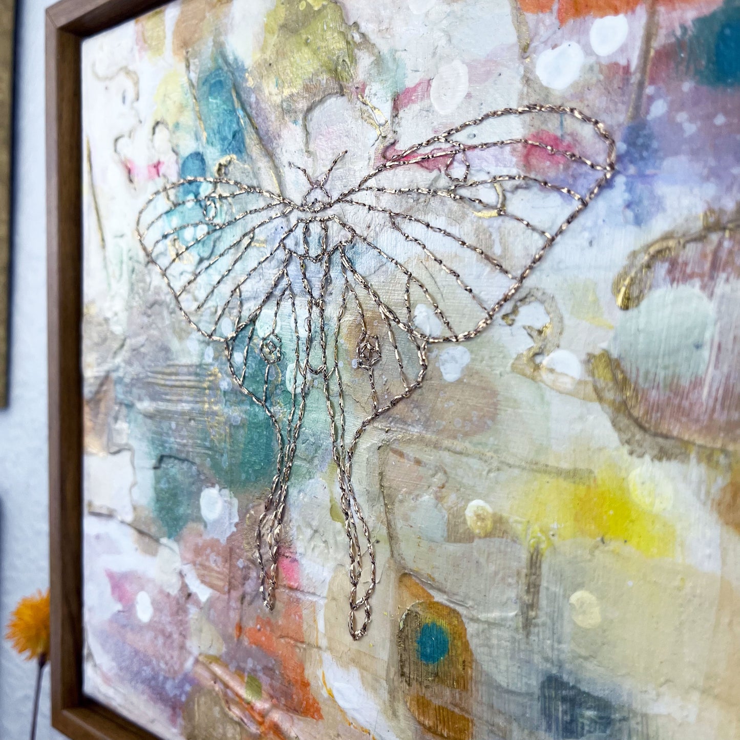 close up angled view of olorful square painting in a wood frame, with a line drawing of a luna moth hand stitched through it in metallic gold thread, hanging on a wall