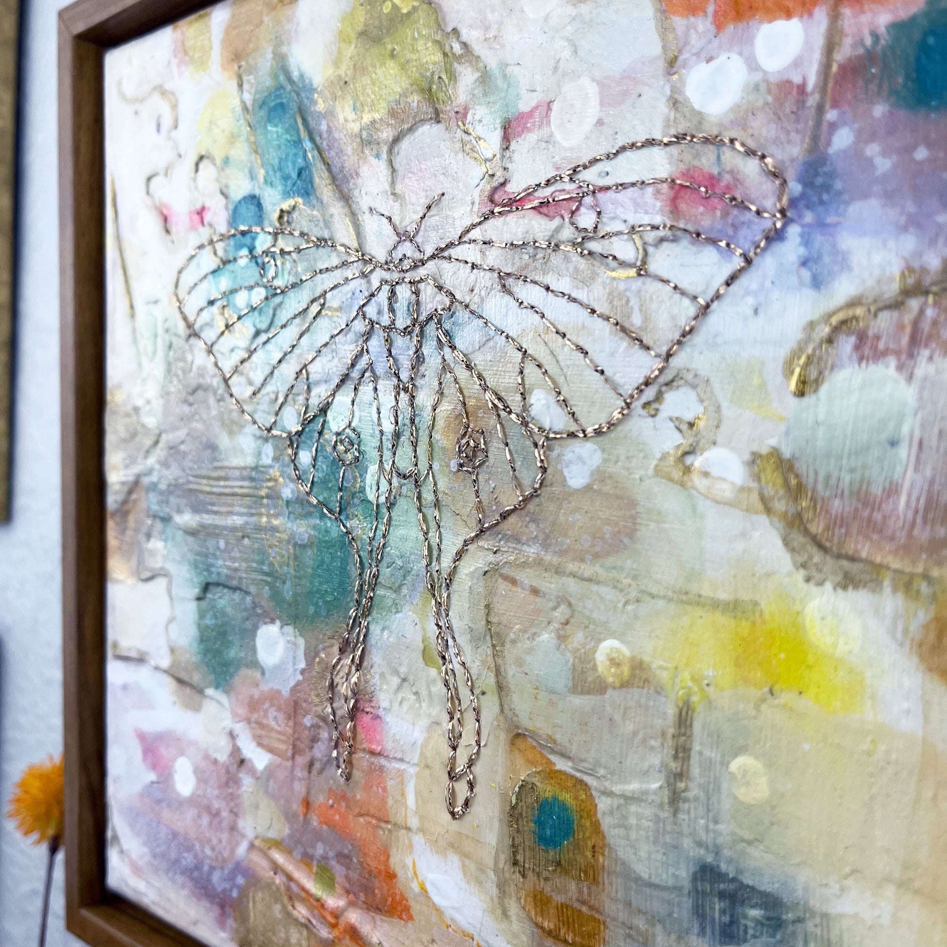 close up angled view of olorful square painting in a wood frame, with a line drawing of a luna moth hand stitched through it in metallic gold thread, hanging on a wall