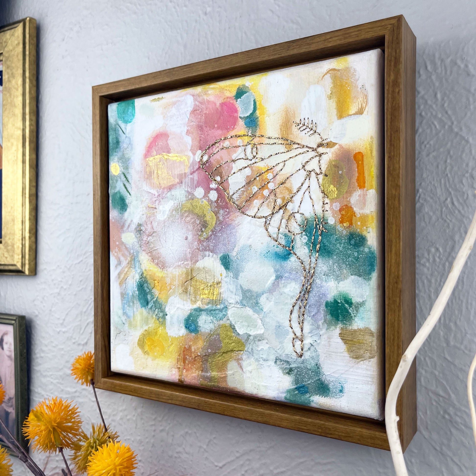 angled view of  a colorful square painting with a hand stitched half of a luna moth in metallic gold thread, in a wood frame hanging on a wall 