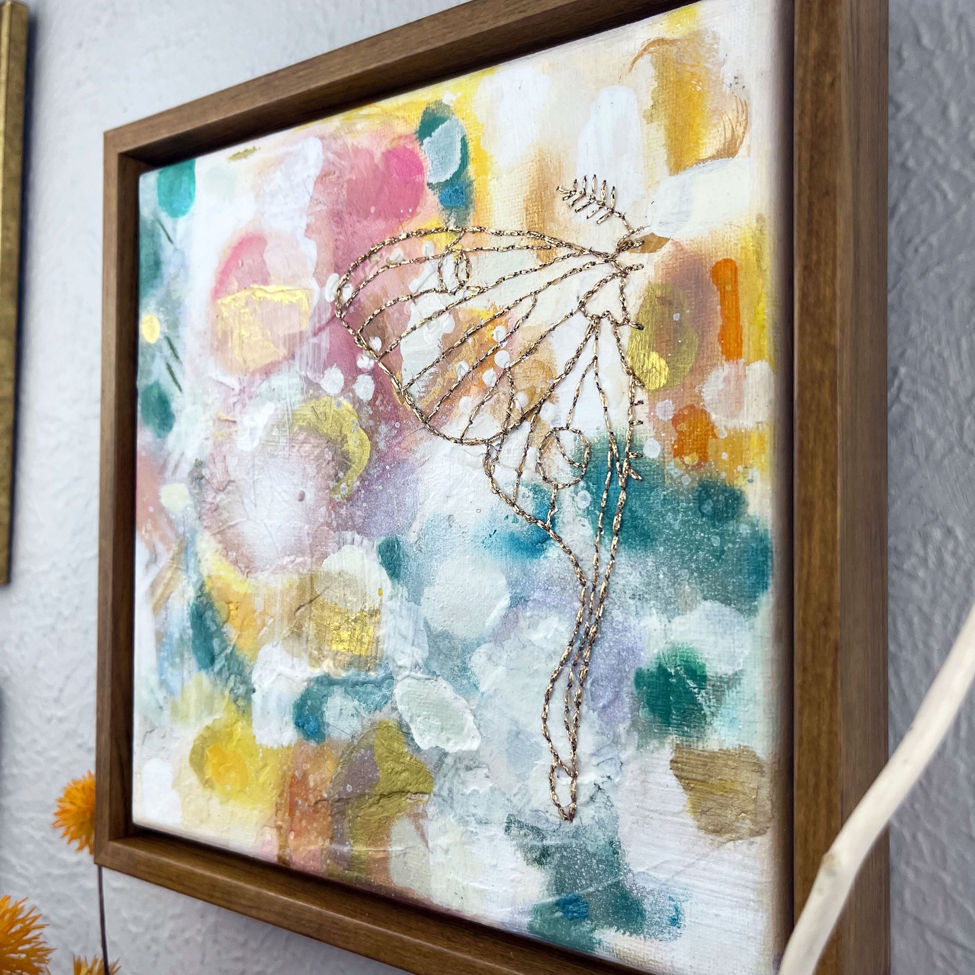 angled view of  a colorful square painting with a hand stitched half of a luna moth in metallic gold thread, in a wood frame hanging on a wall 