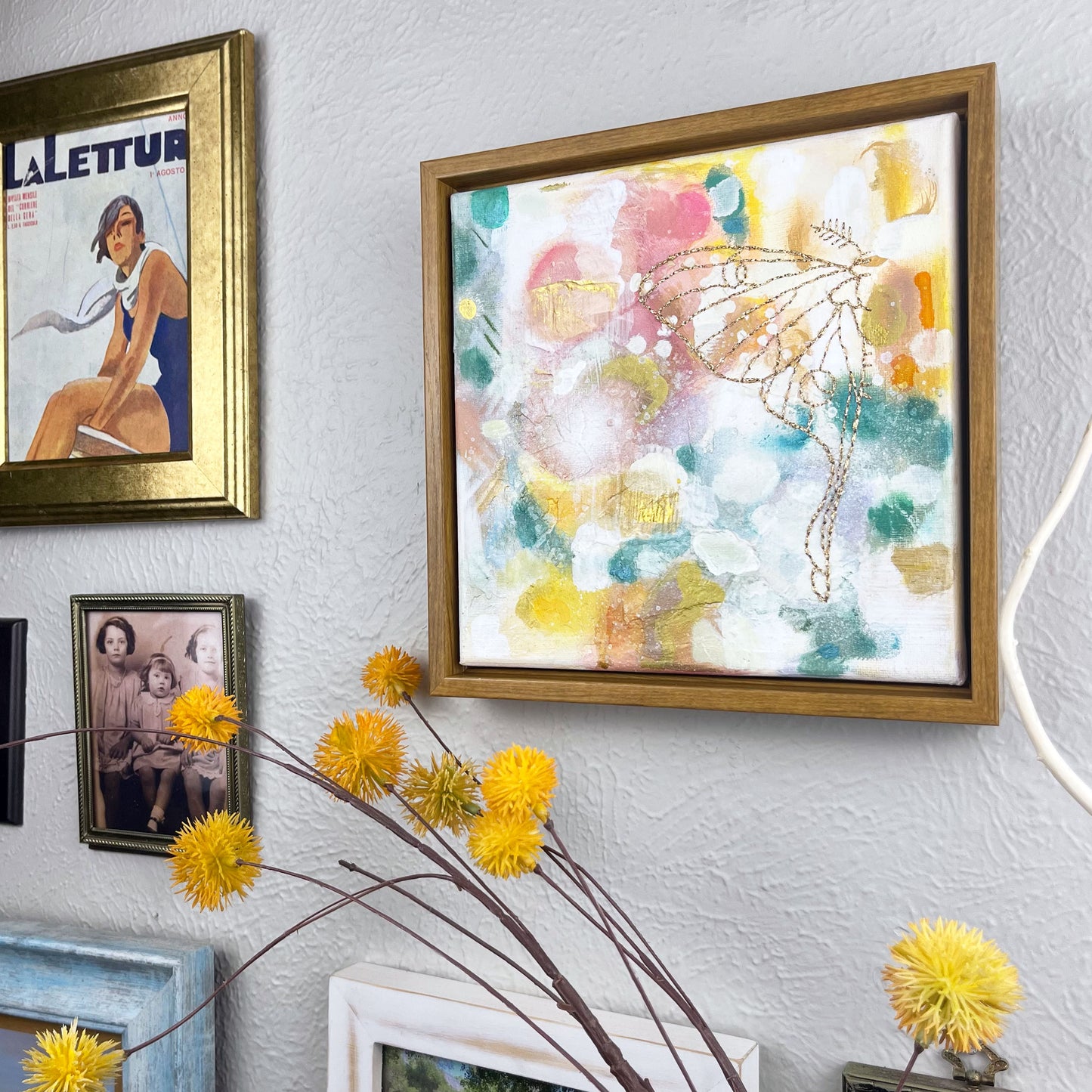  a colorful square painting with a hand stitched half of a luna moth in metallic gold thread, in a wood frame hanging on a wall near other framed art and photos