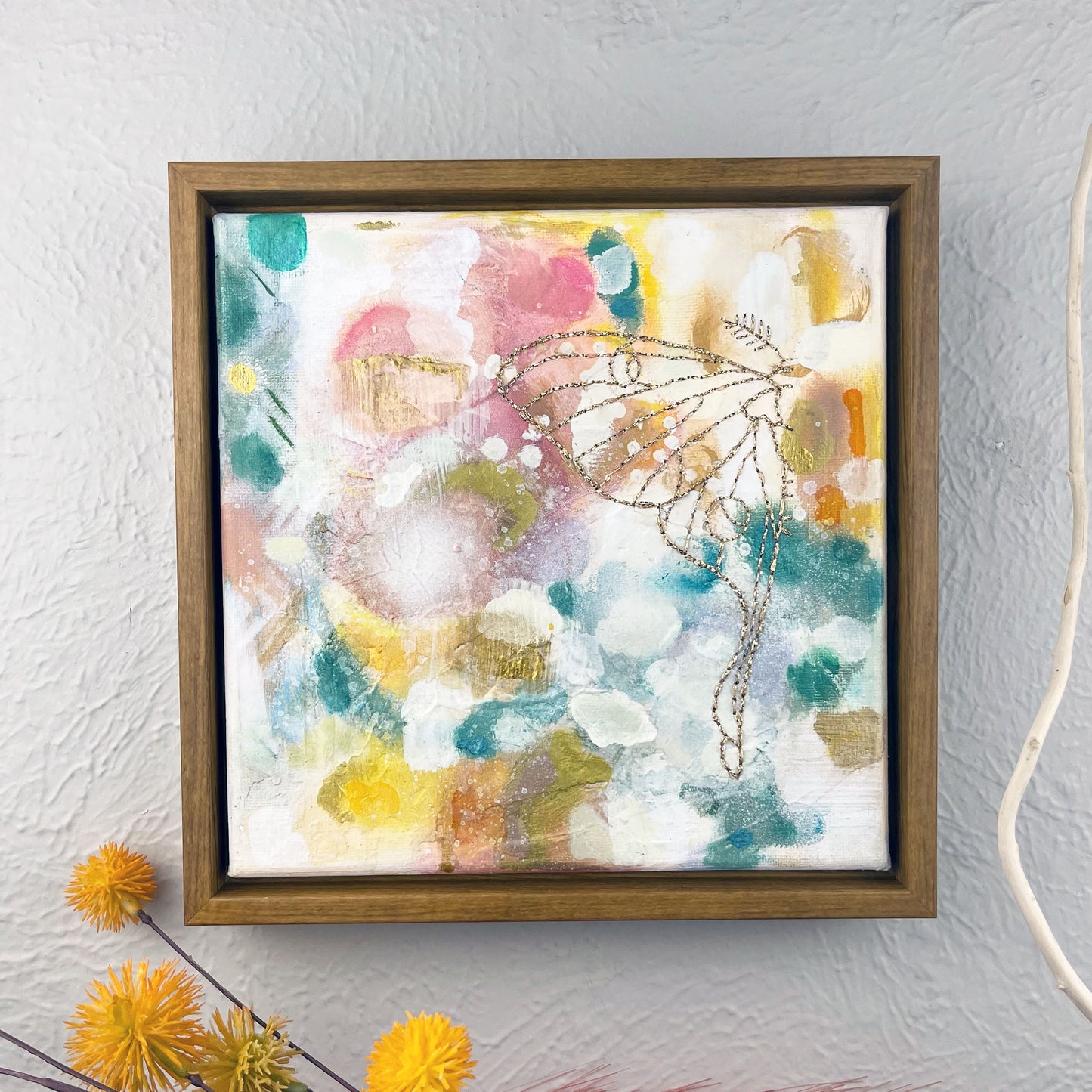 a colorful square painting with a hand stitched half of a luna moth in metallic gold thread, in a wood frame hanging on a wall 