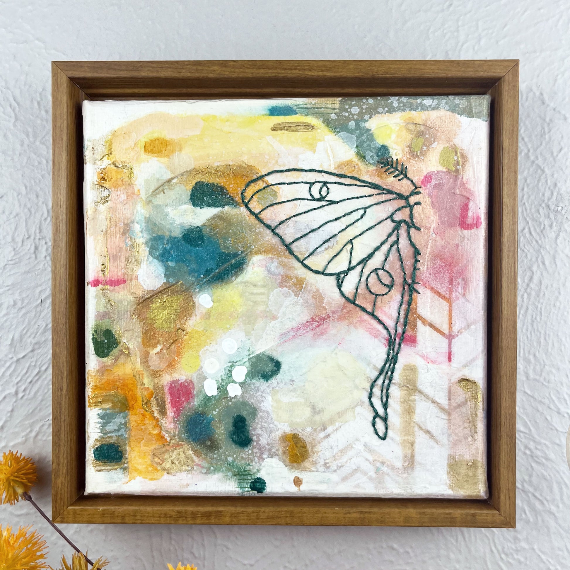 a colorful square painting with a hand stitched half of a luna moth in emerald green embroidery floss, in a wood frame hanging on a wall