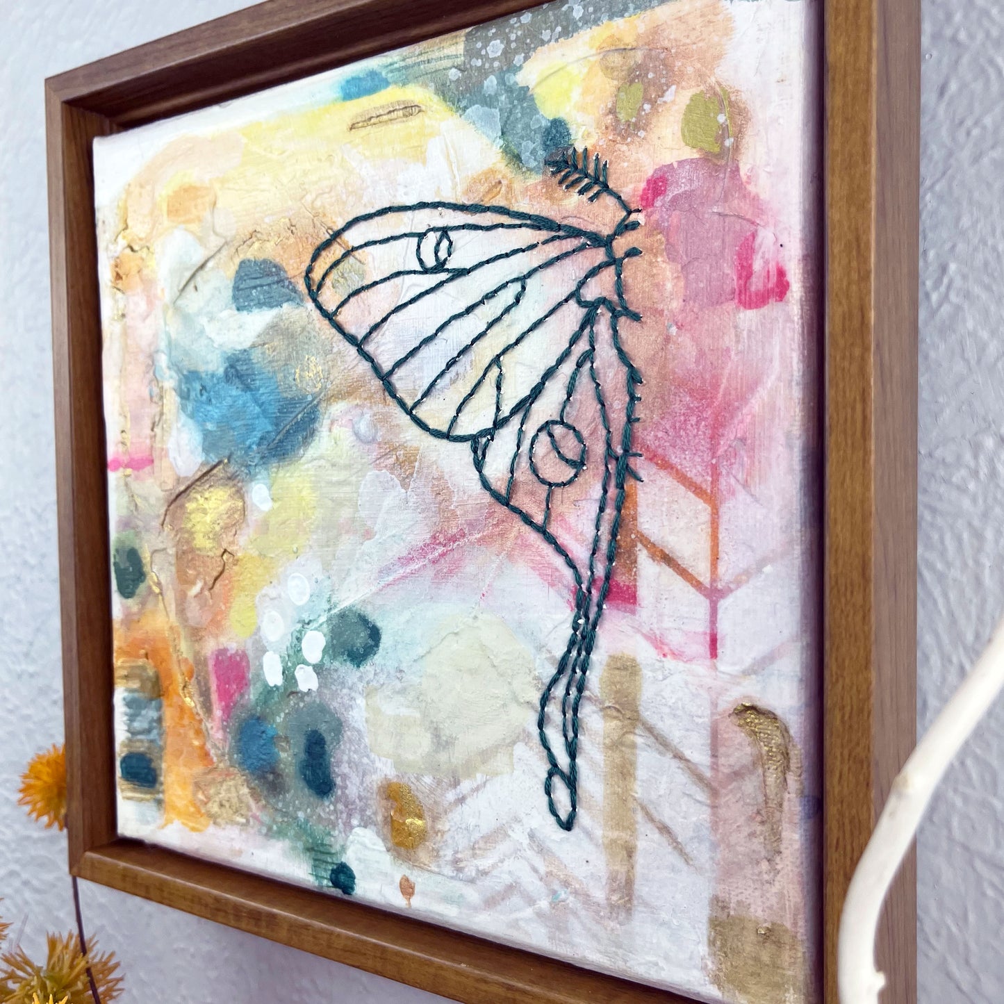 angled view of  a colorful square painting with a hand stitched half of a luna moth in emerald green embroidery floss, in a wood frame hanging on a wall 