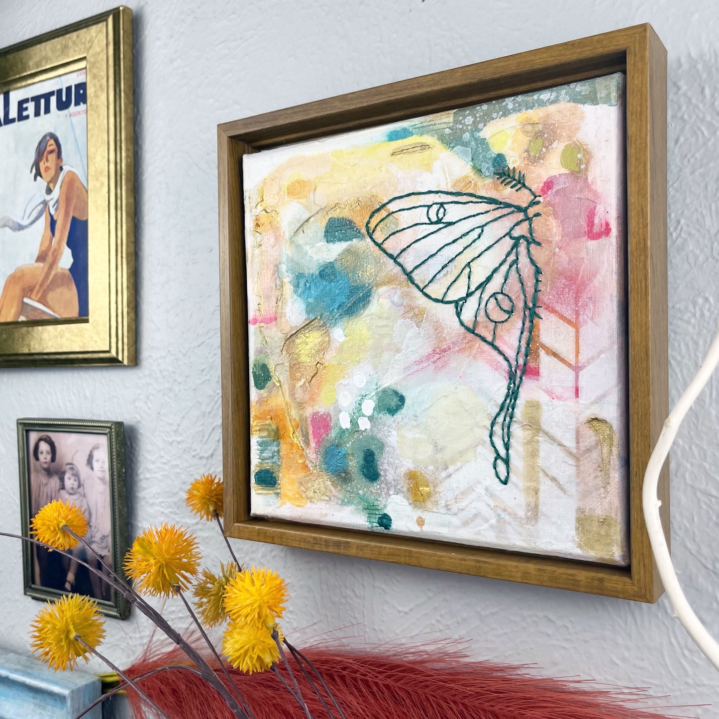 angled view of  a colorful square painting with a hand stitched half of a luna moth in emerald green embroidery floss, in a wood frame hanging on a wall near other framed art and photos
