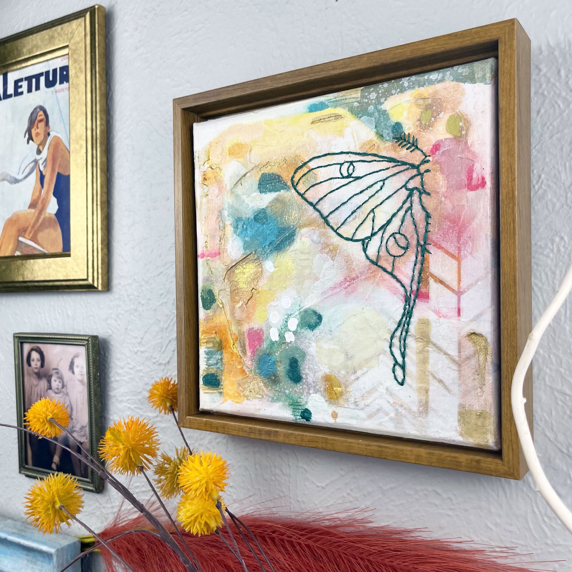 angled view of  a colorful square painting with a hand stitched half of a luna moth in emerald green embroidery floss, in a wood frame hanging on a wall near other framed art and photos