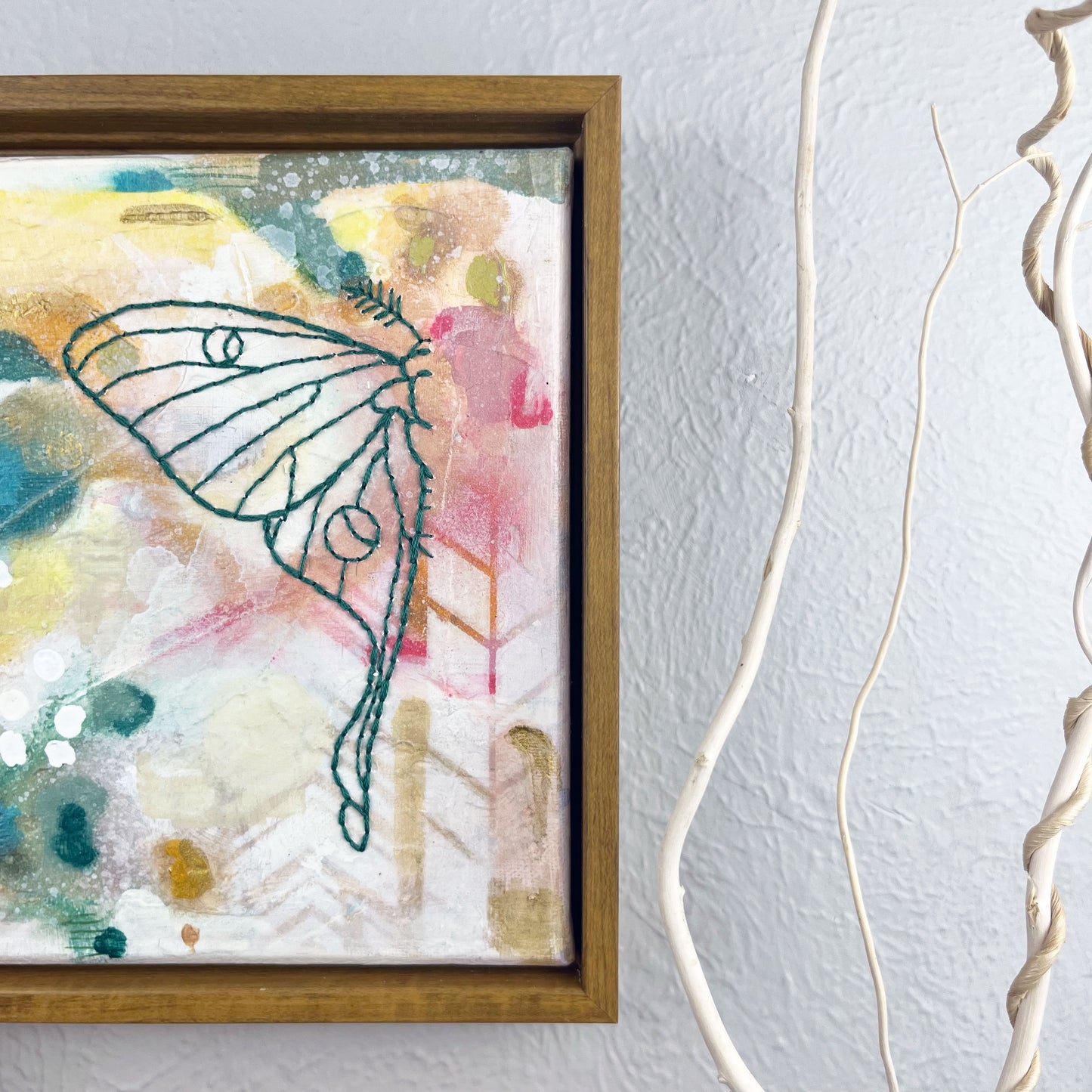 off center view of  a colorful square painting with a hand stitched half of a luna moth in emerald green embroidery floss, in a wood frame hanging on a wall 