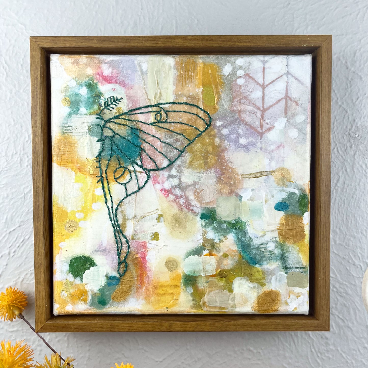 colorful square painting with a hand stitched half of a luna moth in emerald green embroidery floss, in a wood frame hanging on a wall