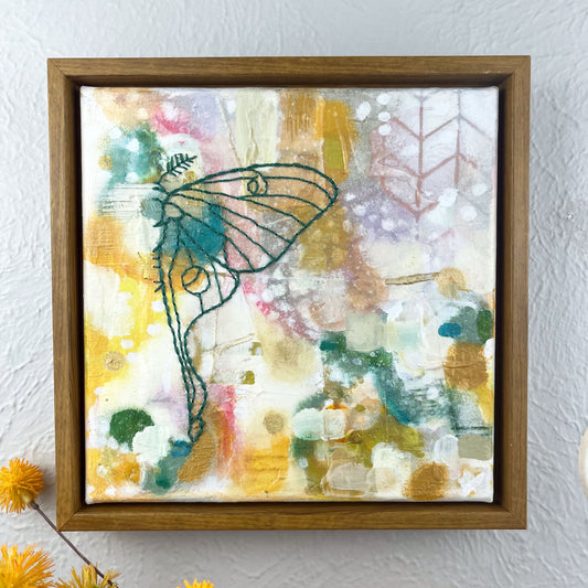 colorful square painting with a hand stitched half of a luna moth in emerald green embroidery floss, in a wood frame hanging on a wall