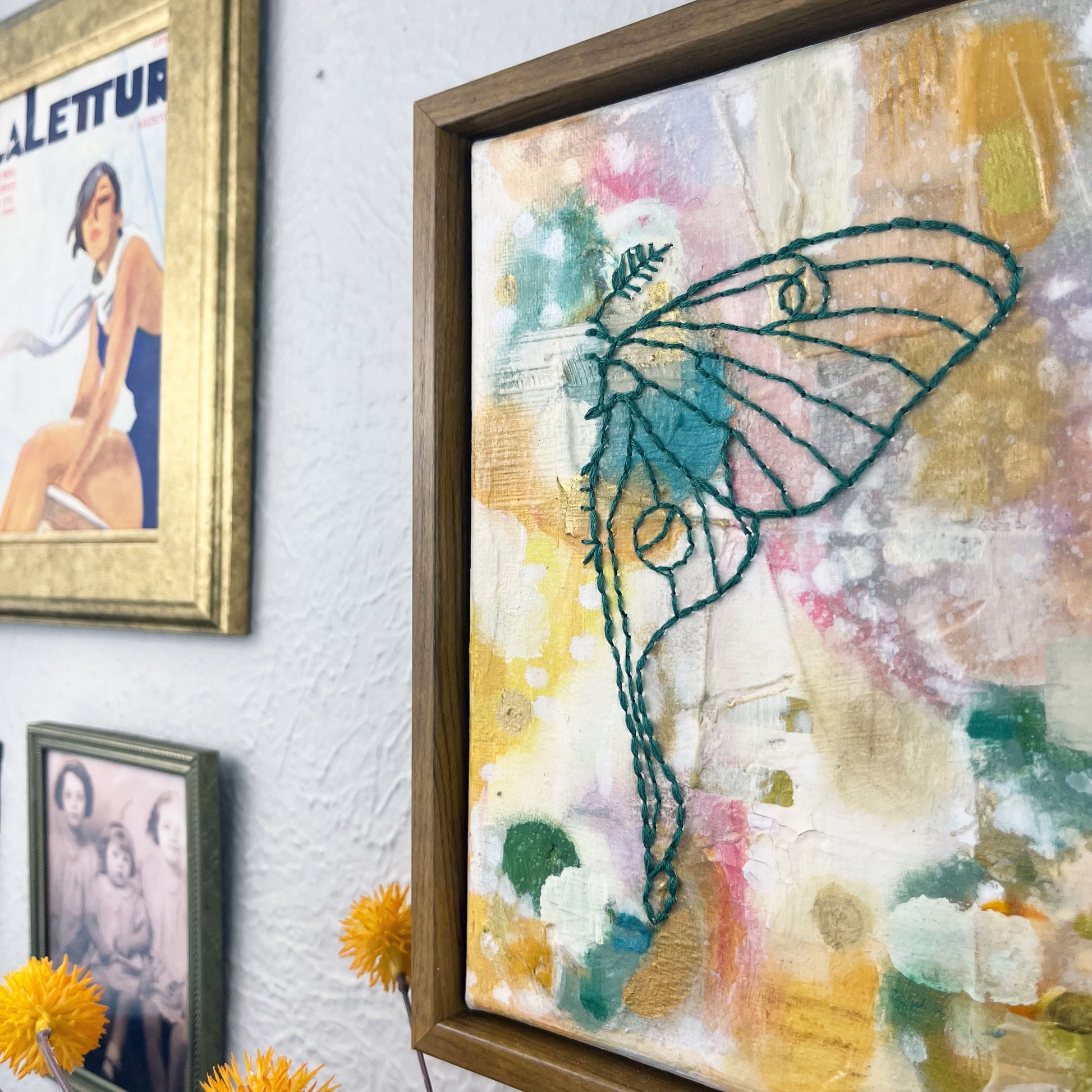 angled view of  a colorful square painting with a hand stitched half of a luna moth in emerald green embroidery floss, in a wood frame hanging on a wall hanging near other framed art and photos