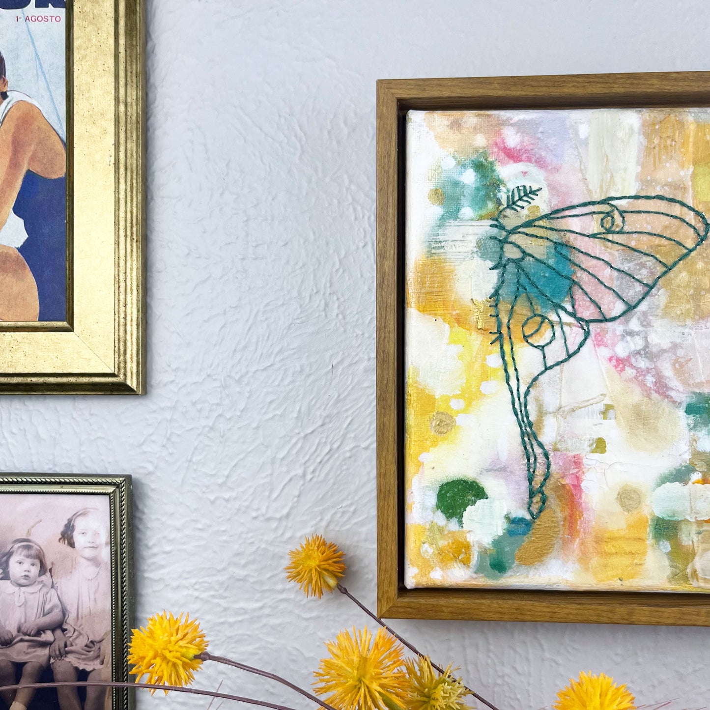 off center view of  a colorful square painting with a hand stitched half of a luna moth in emerald green embroidery floss, in a wood frame hanging on a wall hanging near other framed art and photos