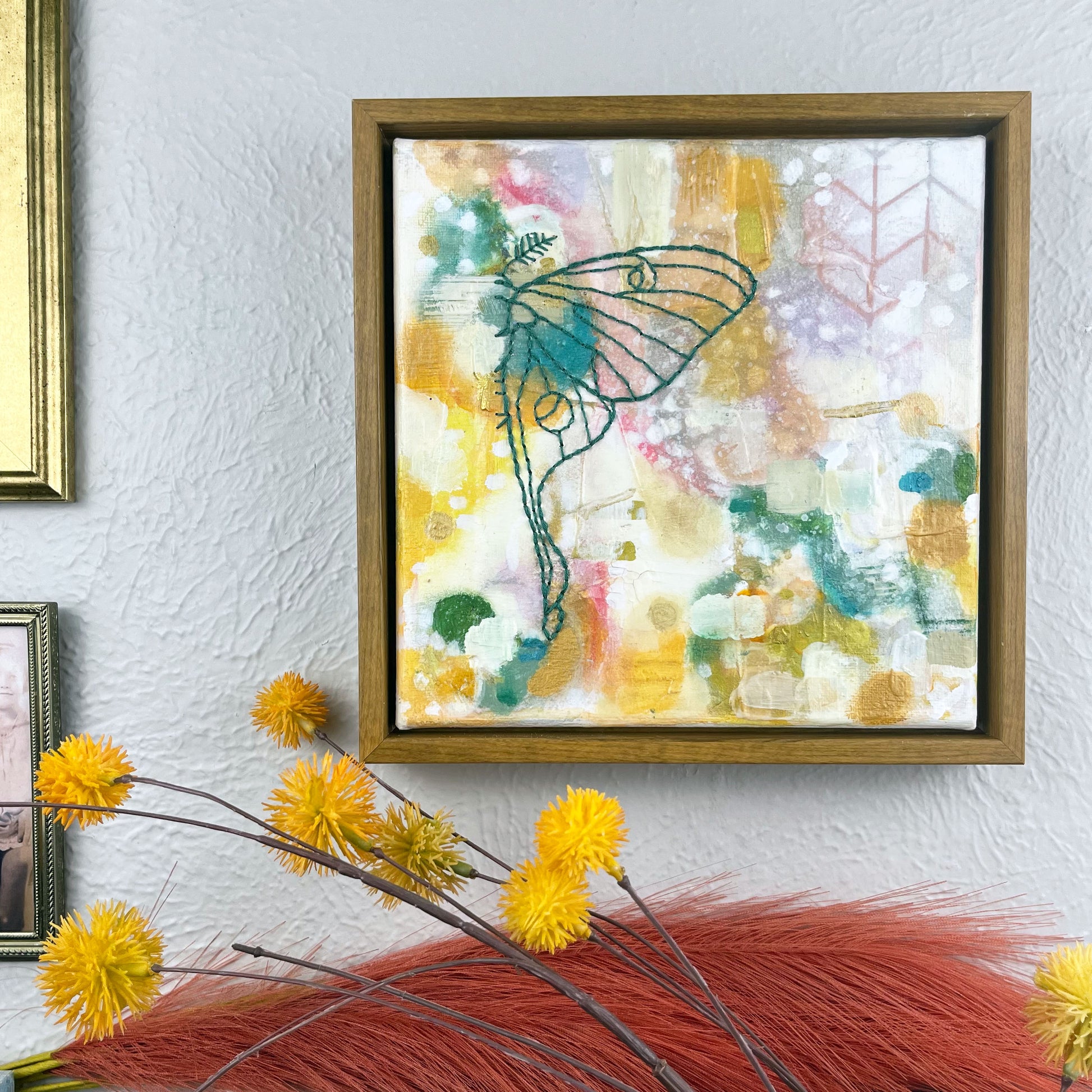 off center view of  a colorful square painting with a hand stitched half of a luna moth in emerald green embroidery floss, in a wood frame hanging on a wall hanging near other framed art and photos