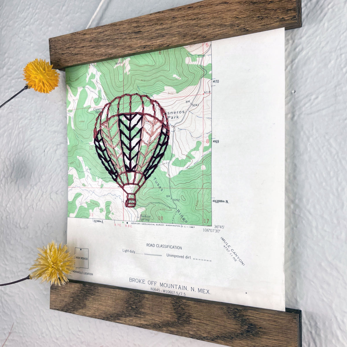 a vintage green and white USGS map of New Mexico hand embroidered with a hot air balloon in shades of mauve, framed on top and bottom with a rustic wood magnetic frame hanging on a grey wall with botanical decorations around it