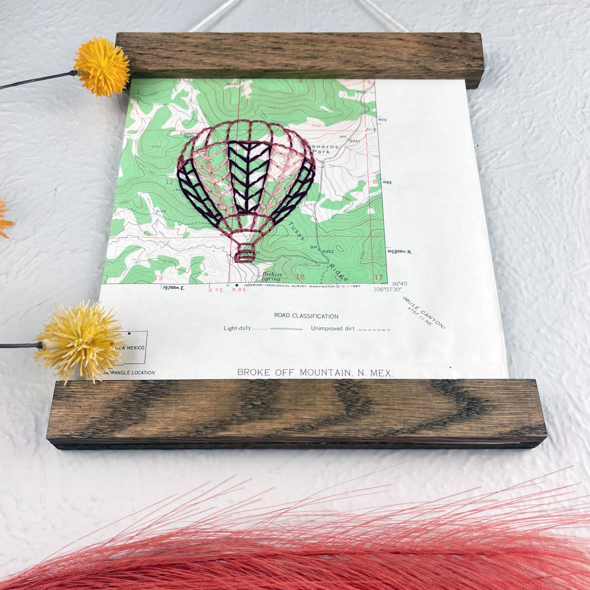 a vintage green and white USGS map of New Mexico hand embroidered with a hot air balloon in shades of mauve, framed on top and bottom with a rustic wood magnetic frame hanging on a grey wall with botanical decorations around it