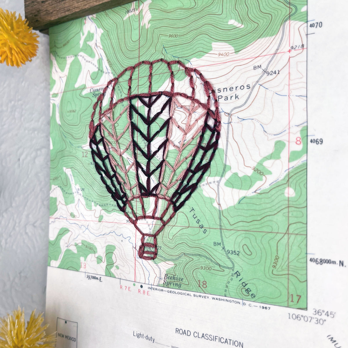 a vintage green and white USGS map of New Mexico hand embroidered with a hot air balloon in shades of mauve, framed on top and bottom with a rustic wood magnetic frame hanging on a grey wall with botanical decorations around it
