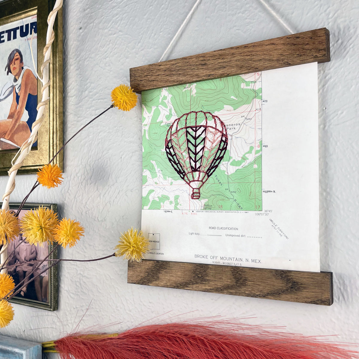 a vintage green and white USGS map of New Mexico hand embroidered with a hot air balloon in shades of mauve, framed on top and bottom with a rustic wood magnetic frame hanging on a grey wall with framed art and botanical decorations around it